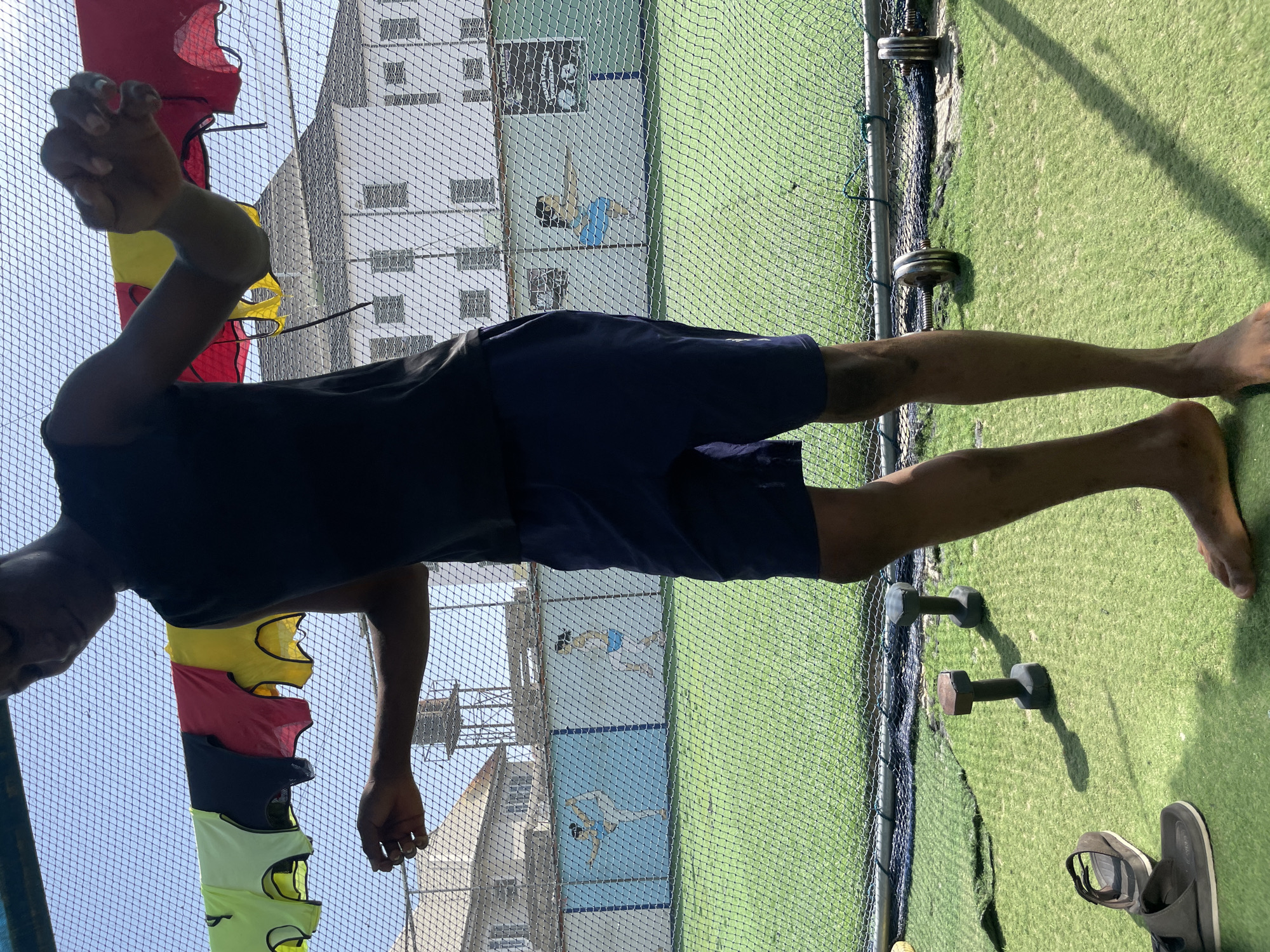 An outdoor scene captured in bright daylight shows a person standing on artificial green turf. The individual, appearing to be male with a dark complexion, is wearing a dark t-shirt and dark shorts, and is barefoot. He stands facing towards the left side of the frame, with his right arm slightly raised and bent, and his left arm relaxed by his side.
In the foreground, near the person's feet, are two dumbbells of different sizes and a pair of dark sandals or flip-flops. Distinct shadows cast by these objects and the person indicate a sunny day, likely midday or early afternoon, with a clear sky visible.
A black mesh net runs vertically across the image, separating the foreground from the background. To the upper left, behind the net, a stack of colorful vests or bibs (red, yellow, black, and light green) hangs vertically. Beyond the net and the vests, a white multi-story building with numerous windows is visible. To the right of the building, also behind the net, is a light blue wall adorned with white stylized figures of people engaged in what appears to be a sport, possibly padel or table tennis. The setting suggests a sports or training facility in Iranla, Nigeria. No discernible text is visible.
