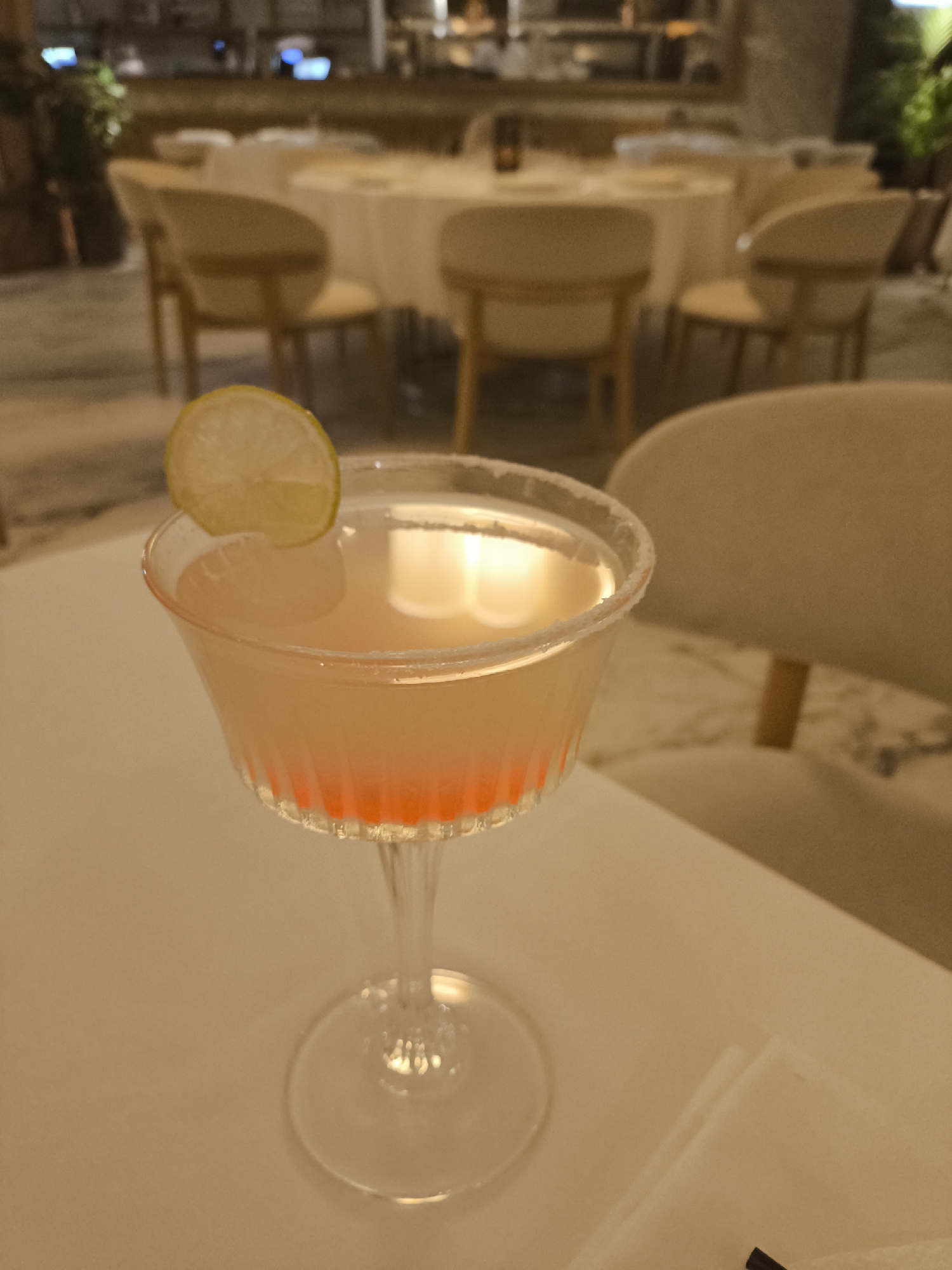 A cocktail, garnished with a lemon slice, sits on a white table in what appears to be a restaurant in an unknown city. The cocktail is served in a stemmed glass with a sugared rim, and there is a visible layer of red liquid at the bottom of the glass. Behind the table are several round tables with tan chairs, indicating a dining environment. In the background, a blurred scene suggests a bar or service area with indistinct people and objects. A stack of white napkins is also on the table. The city is unknown.
