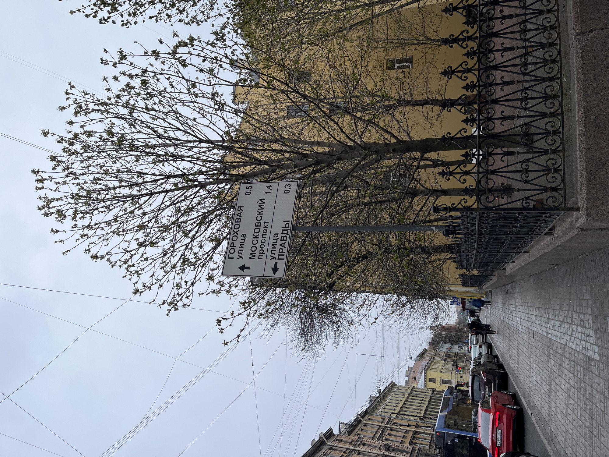 This media file captures an urban street scene on an overcast day, likely in early spring as suggested by the trees with budding leaves. The image is taken from a sidewalk viewpoint, looking down a city street flanked by buildings.
On the right side of the image, a brick-paved sidewalk runs parallel to an ornate black wrought-iron fence. Behind the fence, several trees with slender branches and small green buds rise, partially obscuring a light yellow building.
A white street sign, written in Cyrillic, is mounted on a pole near the center of the image. It indicates directions and distances to local streets:
* "ГОРОХОВАЯ УЛИЦА" (Gorokhovaya Street) - 0.5 km, with an arrow pointing straight ahead/left.
* "МОСКОВСКИЙ ПРОСПЕКТ" (Moskovsky Prospekt) - 1.4 km, with an arrow pointing straight ahead/left.
* "УЛИЦА ПРАВДЫ" (Ulitsa Pravdy) - 0.3 km, with an arrow pointing right.
To the left of the sidewalk and sign, the street is lined with parked vehicles, including a prominent red Audi sedan with a Russian license plate "K 111 УУ 178". The "178" region code on the license plate identifies St. Petersburg, Russia, as the location. Further down the street, a blue bus is visible, along with more parked cars and distant buildings. Several pedestrians can be seen walking on the far sidewalk in the distance.
Above the buildings and street, a network of overhead power lines stretches across a pale, gray sky, indicative of overcast weather. The architectural style of the buildings, combined with the Cyrillic text and license plate, confirms the setting as a Russian city, specifically St. Petersburg. The overall scene is calm, depicting everyday city life.