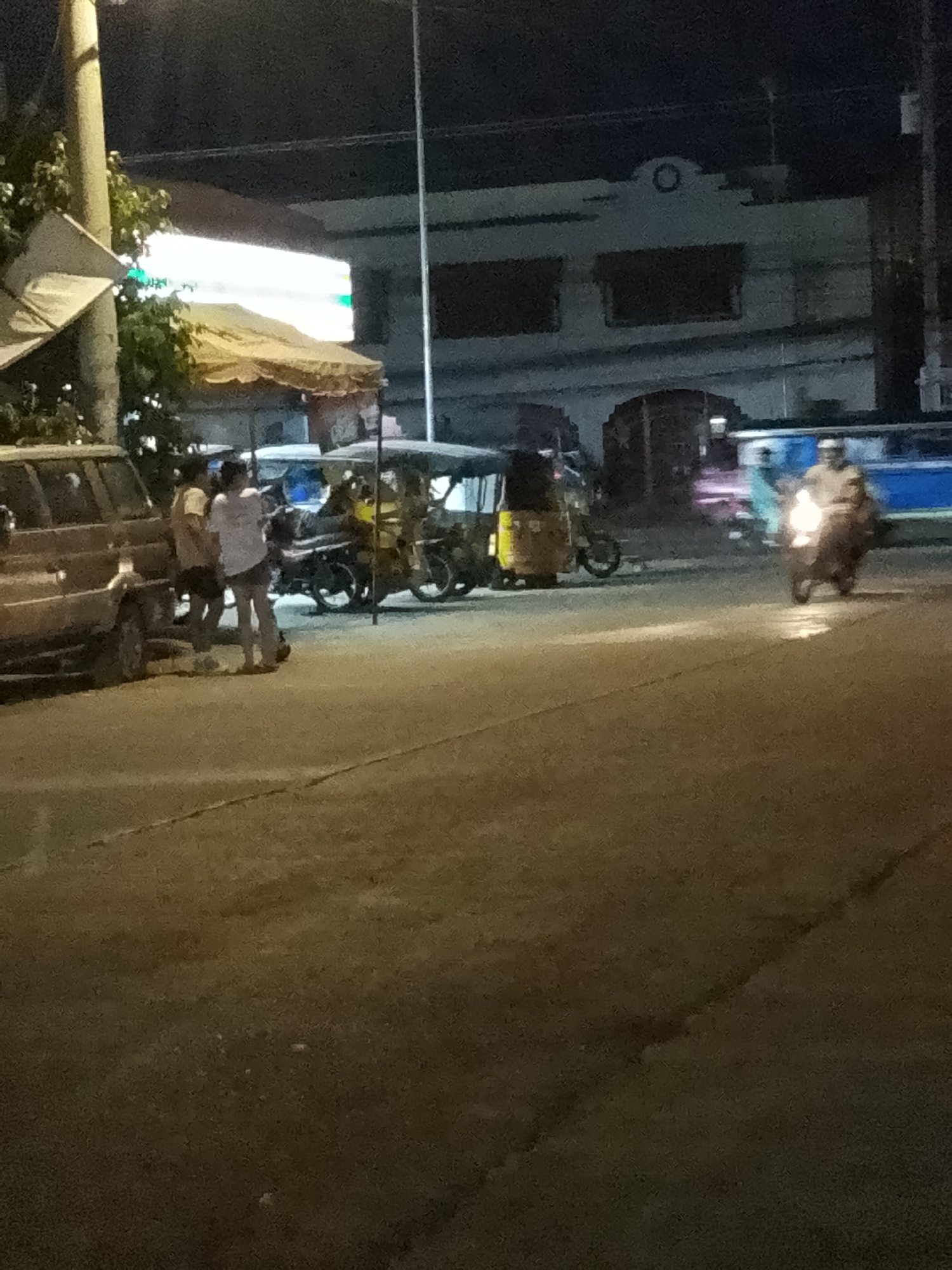 In this nighttime scene, likely in Davao City, Philippines, several people and vehicles are visible on a paved road. The lighting suggests it's after dark, with artificial lights illuminating the area.
In the foreground, to the left, is the side of a dark-colored SUV. Standing near the SUV are three individuals. Two are wearing shorts and t-shirts, and the third person is facing away from the camera. Their posture suggests they are casually gathered or conversing.
Further into the scene, several tricycles are parked or moving. One prominent yellow tricycle with a covered seating area is visible. Another tricycle is behind it, partially obscured. In the background, a blue and white vehicle, possibly a bus or another large vehicle, is present.
To the right of the frame, a person is riding a motorcycle, illuminated by its bright headlight. The rider is wearing a helmet. The road surface shows some texture and possibly some slight wear, with a faint line marking across it.
Behind the vehicles, a building with arched windows and a decorative circular element on its facade can be seen. There's a bright, horizontal illuminated sign above some structures in the mid-ground, possibly indicating a business or advertisement. The overall atmosphere appears to be that of a typical street scene at night in a populated area, with a mix of transportation and pedestrian activity. No text is clearly legible in the image.