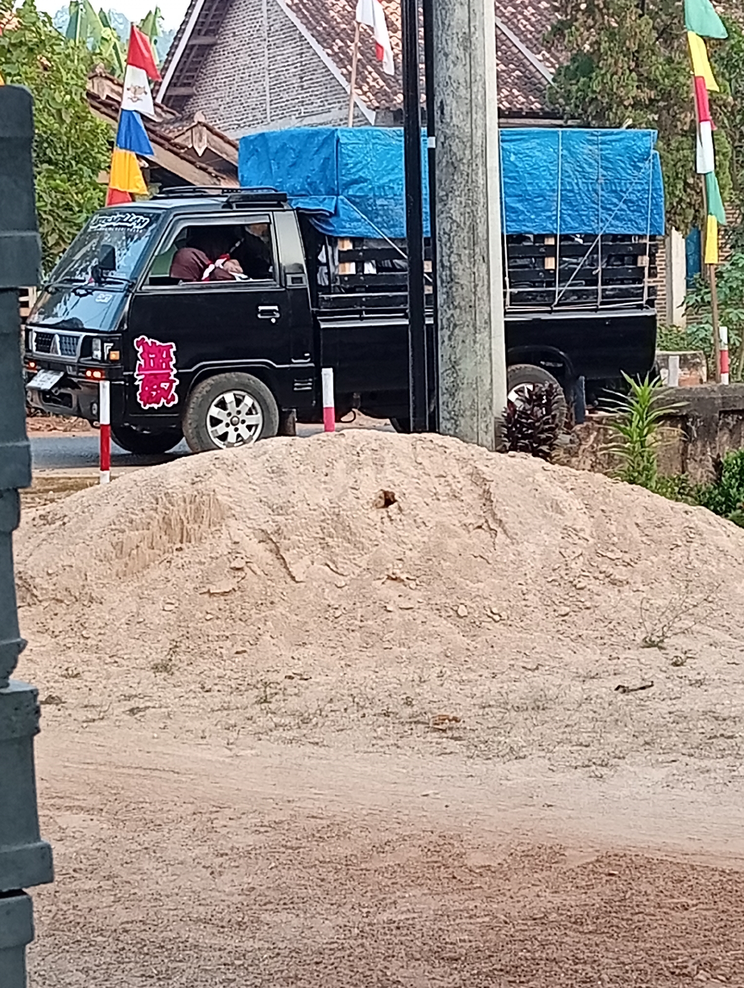 The image shows a black pickup truck parked next to a large pile of sand. The truck has a blue tarp covering its cargo bed. There are several flags with red, white, blue, and yellow colors hanging from poles in the background. A person is visible inside the truck's cabin, appearing to be resting. The scene appears to be on a street with some greenery and buildings in the distance. It is likely in Banyuwangi, Indonesia given the cultural context of the flags and the type of vehicle.