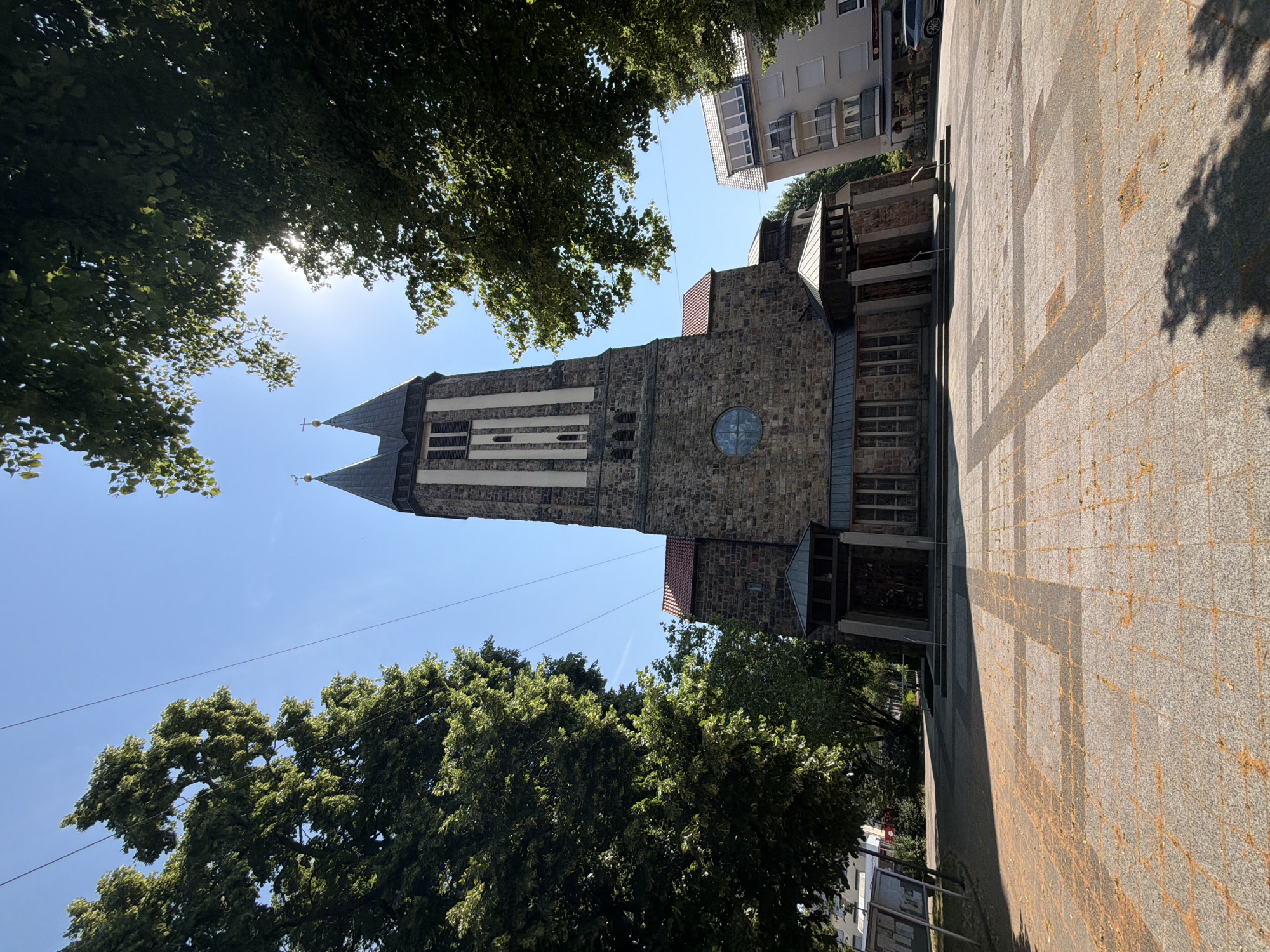 A low-angle shot captures a stone church with two tall spires against a bright blue sky, framed by lush green trees. The church features a large circular stained-glass window and a prominent entrance with a covered porch. The foreground is a paved plaza with a geometric pattern of light and dark stones, lightly scattered with fallen leaves. To the right of the church, a modern apartment building is visible, and a car is parked on the street. The scene appears to be a public space in a city, bathed in sunlight on a clear day.