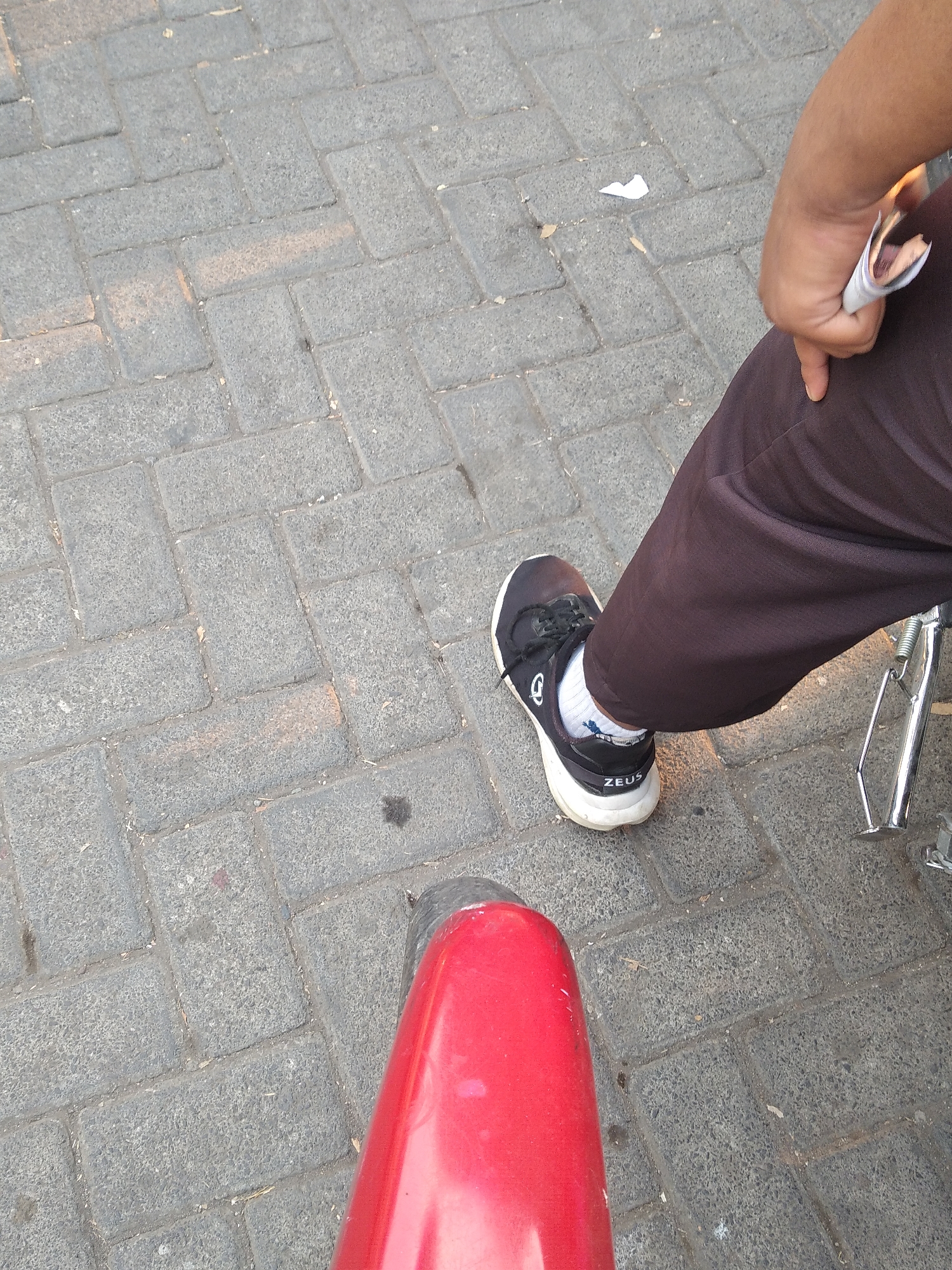 The image shows a close-up view of a person's legs and feet, standing on a paved surface. The pavement is made of interlocking gray bricks with visible grout lines and some scattered debris.
The person is wearing dark brown pants and white socks with black and white athletic shoes. The shoes have "ZEUS" written on the heel. A portion of a shiny metal object, possibly part of a bicycle or motorcycle kickstand, is visible to the right of the person's leg. In the upper right corner, a hand is holding a folded piece of paper.
The scene is set on a street or walkway in Jubung, Indonesia. The lighting suggests it is daytime, with some shadows cast on the pavement. The overall impression is a mundane, everyday scene in an urban or semi-urban environment.