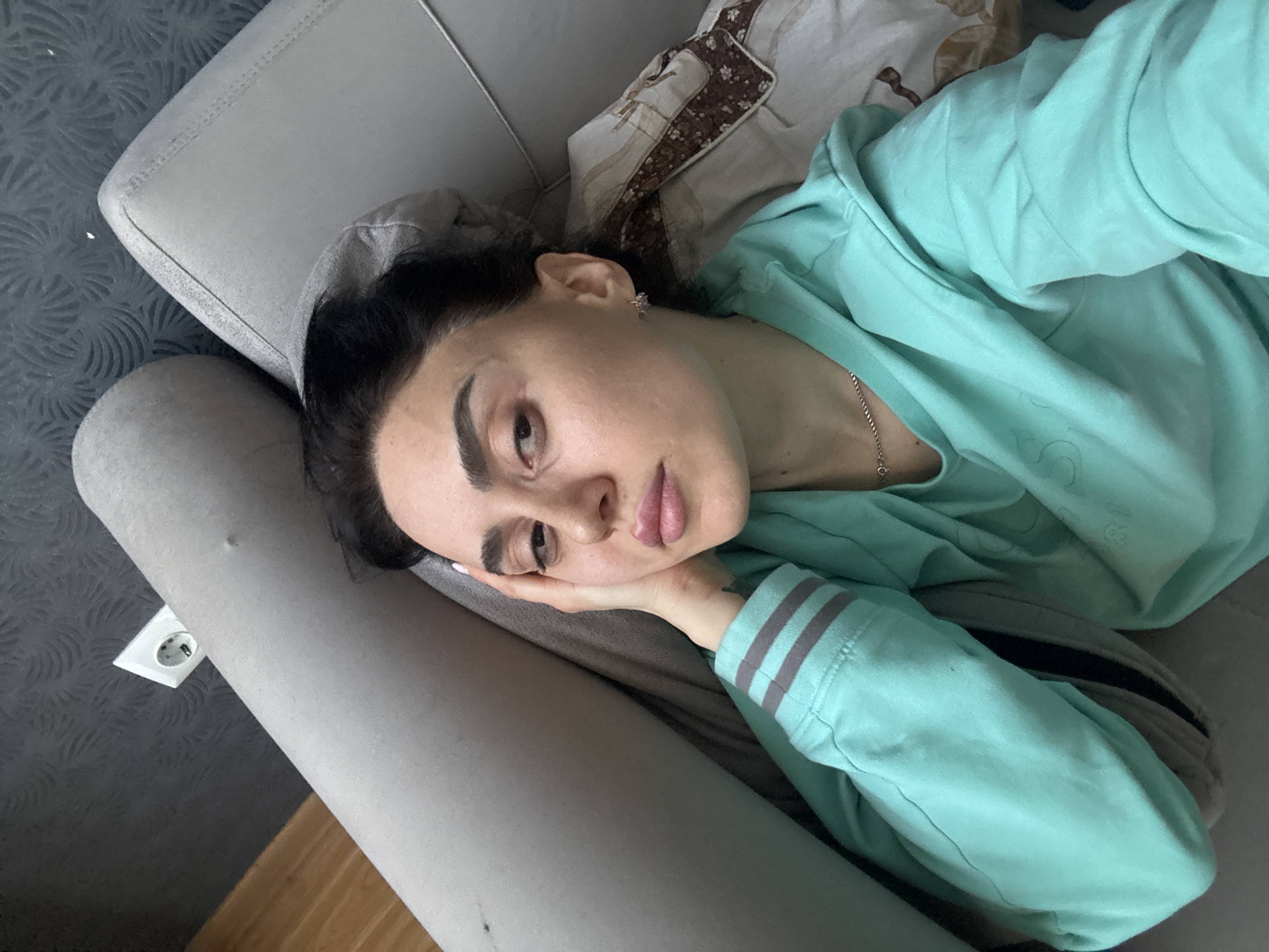 A woman is lying on a gray couch, wearing a light green sweatshirt with gray stripes on the sleeve. She rests her head on her hand and looks toward the camera. A patterned pillow is behind her head. An electrical outlet is visible on the wall near the couch. The location is likely indoors, potentially in a residential setting within Tbilisi, Georgia, based on the electrical outlet type.