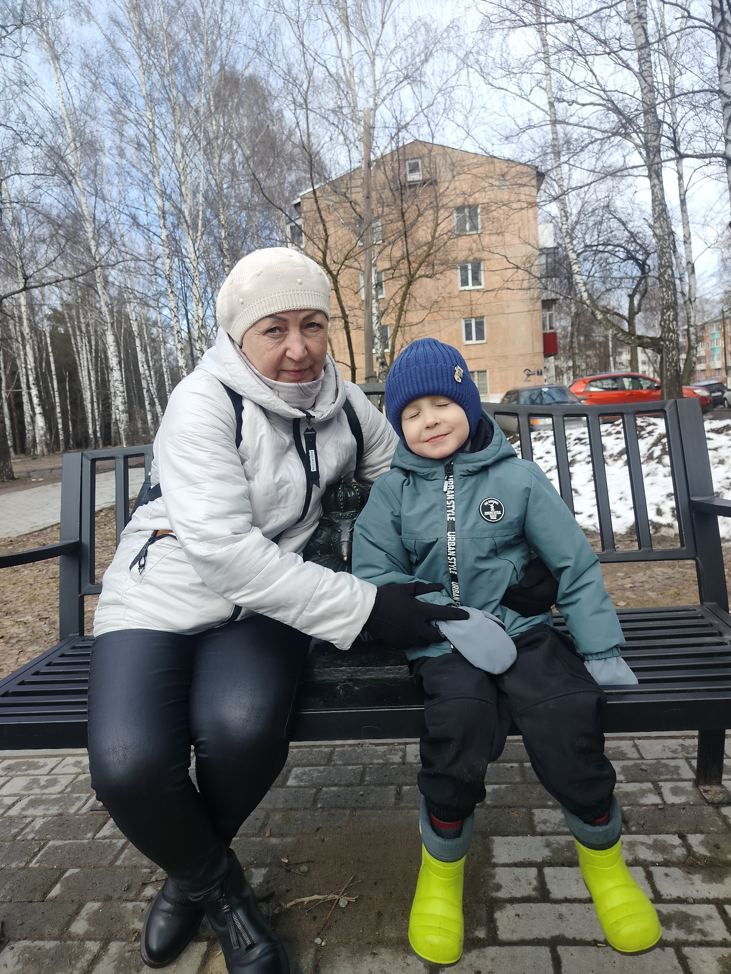 In a city park in Unknown, Unknown, an older woman and a young boy are seated on a black metal bench. The woman, wearing a white quilted jacket, black pants, and black boots, also has on a cream-colored beret. She has her arm around the boy. The boy, wearing a blue beanie, a teal jacket with white lettering, black pants, gray mittens, and bright yellow boots, has his eyes closed. Behind them, there is a backdrop of bare trees, a multi-story apartment building, and a red car parked on the street. The ground appears to be paved with gray bricks or stones.
