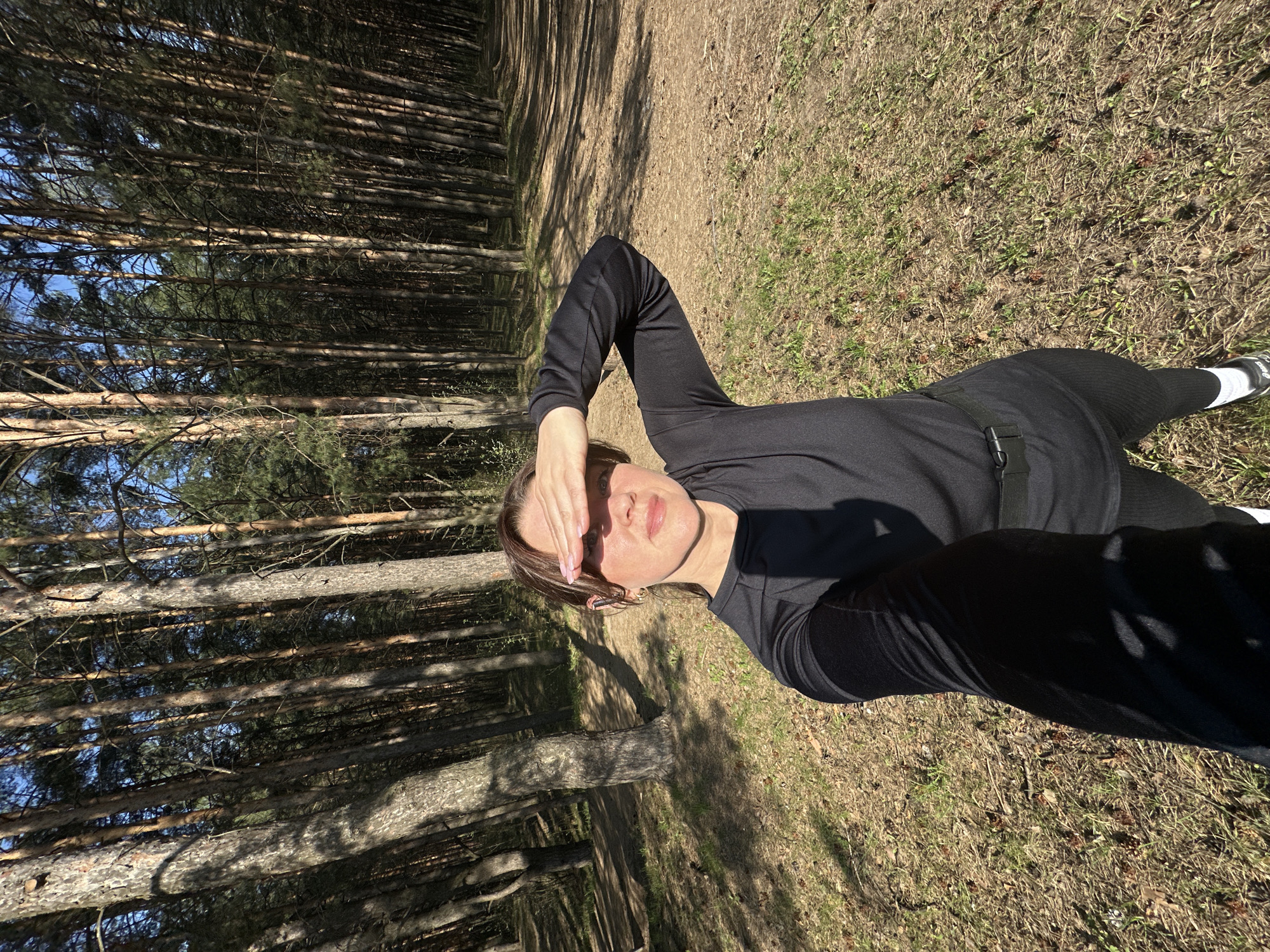 A woman is lying on the ground in a forest in Chaikovsky, Russia, with her arm raised to shield her eyes from the sun. She is wearing black clothing. The forest floor is covered in dry grass and pine needles. Tall pine trees line the pathway. The sky is visible through the trees, showing a clear blue sky. The photo has been taken at an angle, with the camera tilted to the side.