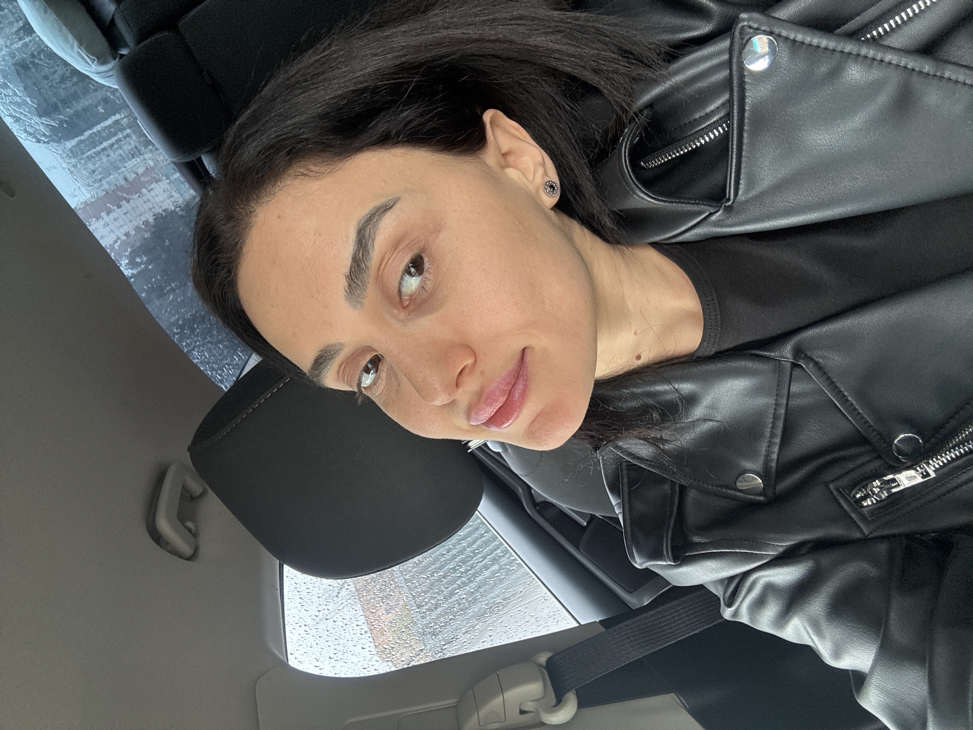 A woman with dark hair and brown eyes is in the passenger seat of a car. She is wearing a black leather jacket. Raindrops are visible on the car window. Based on the rain and architecture visible through the window, the location is consistent with the city of Tbilisi, Georgia. The seatbelt is buckled.