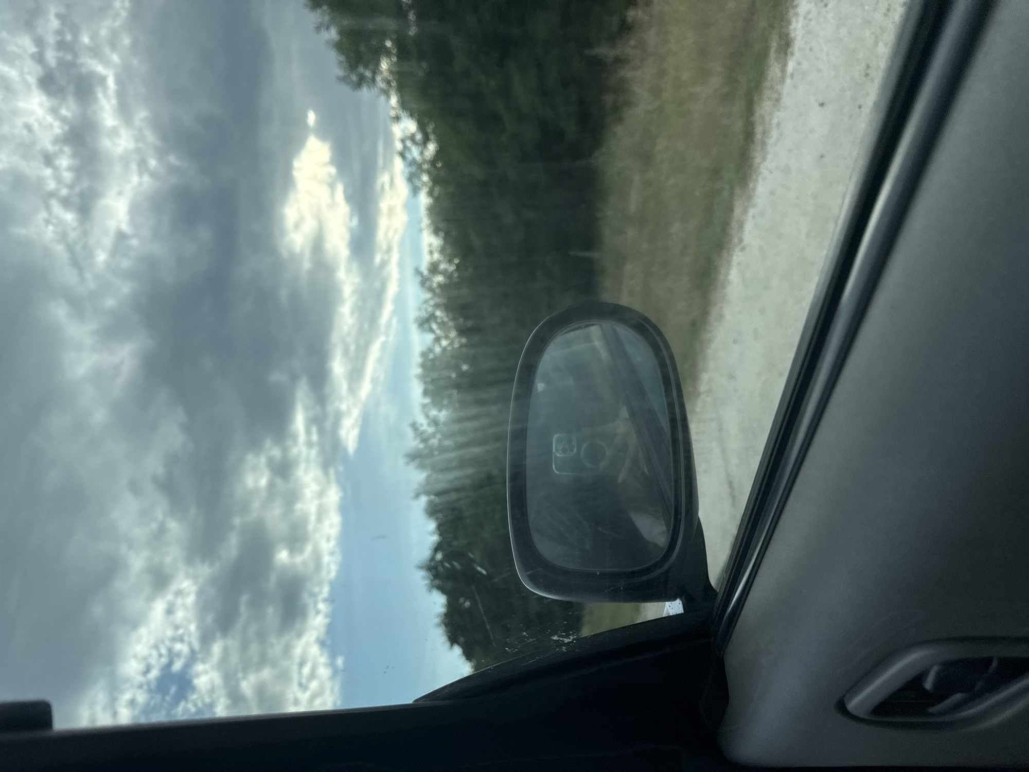 The image captures a view from inside a car, looking out the side window. The car's dark door panel occupies the foreground on the left. A side-view mirror is visible, reflecting a person holding a smartphone, presumably taking the photo. Beyond the mirror, the landscape appears rural, with a dirt or gravel road running alongside a dense line of green trees. The sky is filled with dramatic, cloudy formations, with patches of sunlight breaking through. The specific location in Razeni, Moldova, is not discernible from the visual information provided in the image.