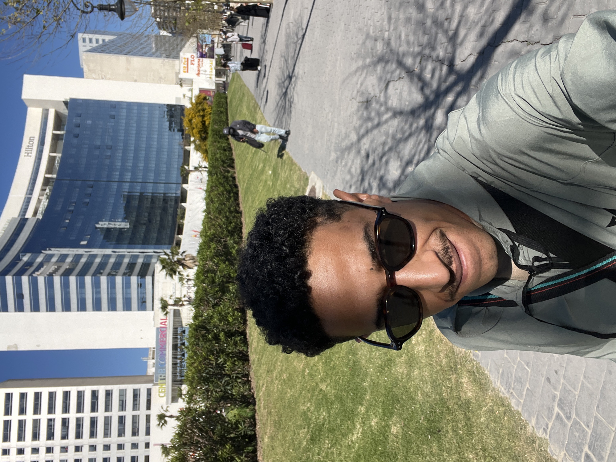The image depicts an outdoor, daytime scene in Tanger, Morocco, specifically in front of the Hilton Tanger City Center and the adjacent Tanger City Center commercial complex. The foreground is dominated by a male individual in a close-up, selfie-style shot. He has dark, curly hair, a light mustache, and wears dark sunglasses, a light grey-green long-sleeved jacket, and a dark shoulder strap with blue, black, and red/orange stripes. His expression is a slight smile.
To the subject's left is a well-maintained green lawn bordered by a hedge. To his right, a grey paved walkway extends into the mid-ground. Overhead, a clear blue sky is visible. In the background, modern high-rise buildings are prominent. The Hilton Tanger City Center, with its curved, dark glass facade and "Hilton" signage, is clearly visible, along with the white high-rise structure of the Tanger City Center featuring "CENTRE COMMERCIAL" signage. Palm trees and other foliage are interspersed around the buildings.
Several pedestrians are visible on the paved walkway in the mid-ground. One individual, wearing a dark jacket, light jeans, and a baseball cap, walks leftward, carrying a dark bag over their shoulder. Further down the walkway, additional pedestrians are present, some with rolling luggage, including a red and a black suitcase. Strong sunlight illuminates the scene, casting tree shadows onto the paved ground.