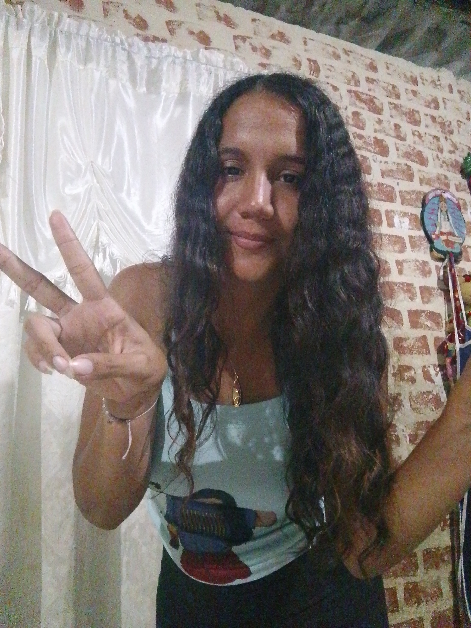 A medium shot depicts a woman with long, wavy dark hair and light skin giving the peace sign. She is wearing a light blue tank top with a cartoon design and black pants. A gold necklace and silver bracelet adorn her.
Behind her, a backdrop of white, slightly textured curtains hangs to the left, while a brick wall stretches to the right. A circular object with an image is affixed to the wall to the right of her head.
The location in the city of Juan Griego, Venezuela, is assumed and is not verifiable from the image. The image could have been taken anywhere.