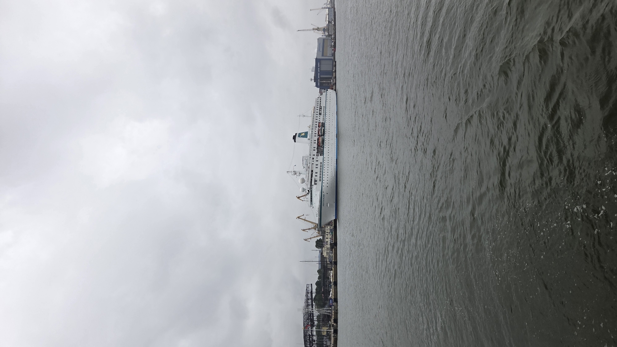 An overcast day in the city of Klaipėda, Lithuania, features a large white cruise ship, the MS Deutschland, docked in the port. The ship, with its name clearly visible on the hull, is the central focus, positioned in the middle ground of the image. Behind the ship, the industrial port area is visible, characterized by cranes, warehouses, and other port infrastructure. To the left, a large, dark, industrial structure with intricate scaffolding dominates the background. The water in the foreground is choppy and dark, reflecting the grey sky. The overall scene is one of maritime activity under a dull, cloudy sky.