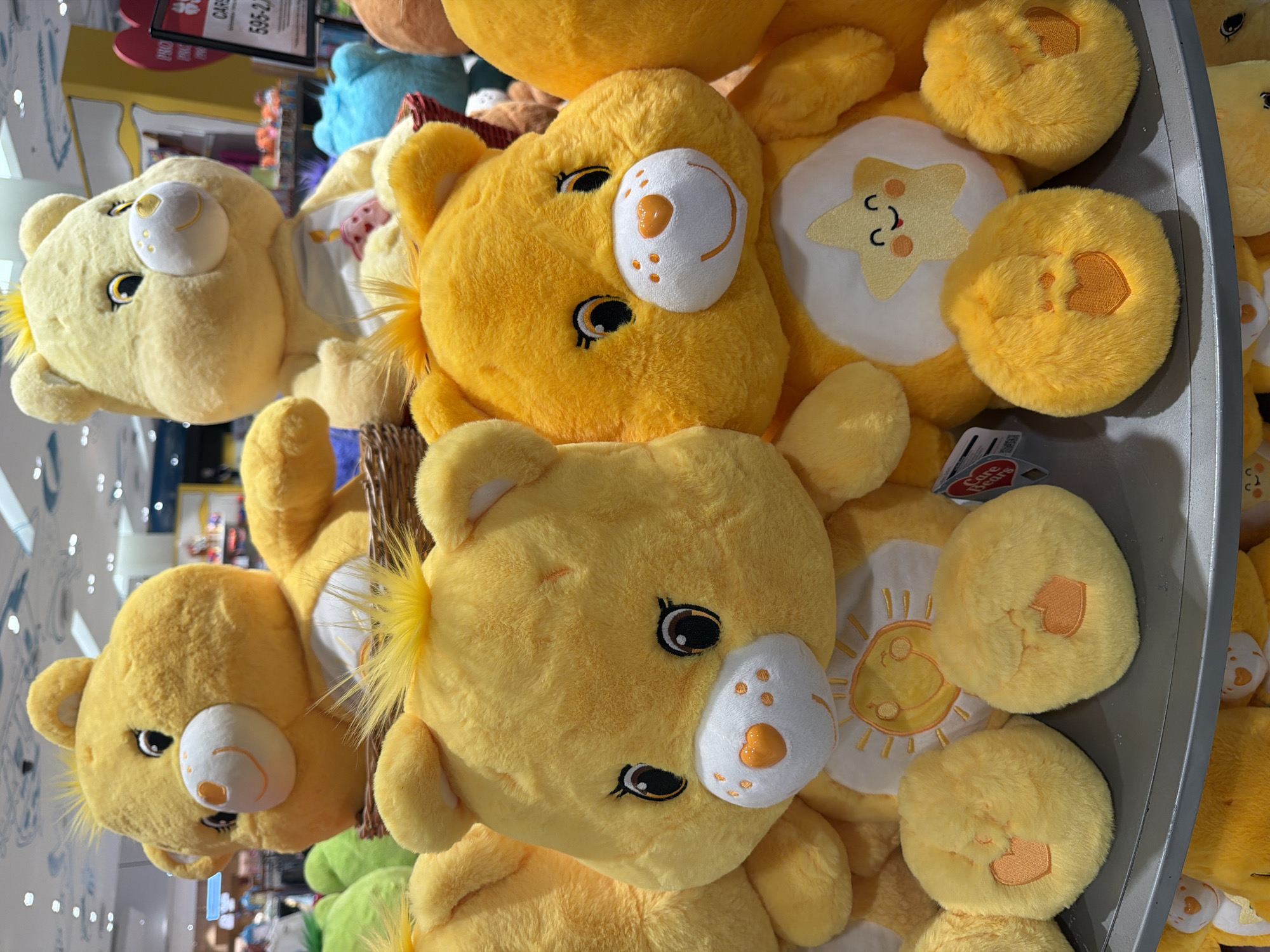 The image displays a collection of yellow Care Bear stuffed toys arranged on display shelves. The foreground features two prominent yellow Care Bears, one with a sun symbol on its chest and the other with a star symbol. Their paws, embroidered with heart shapes, are visible at the bottom of the frame. Behind them, other Care Bears, including a lighter yellow one and a blue one, are partially visible. The background suggests an indoor retail environment, possibly a toy store, with shelves stocked with merchandise. There is no indication of the city of Ko Samui District, Thailand, in the image itself. The scene is static, with no visible people or actions.