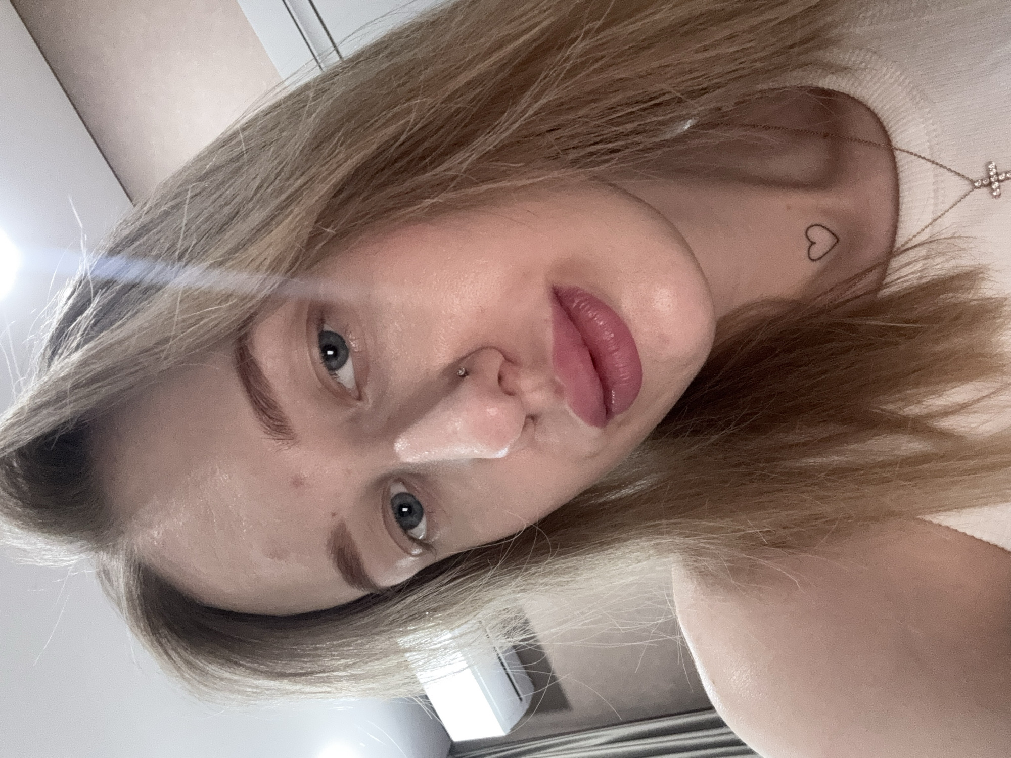 A close-up, selfie-style photograph shows a fair-skinned young woman with blonde hair, blue eyes, and a heart tattoo on her neck. Her lips are slightly parted, and a small amount of white substance is visible on her nose. She has a piercing on the left side of her nose. She is wearing a white top and a delicate necklace with a cross pendant is visible. The background is blurry, appearing to be an interior setting with light walls and a glimpse of a radiator. The image is not clear enough to determine a specific location in Ovoshchnoy, Russia.