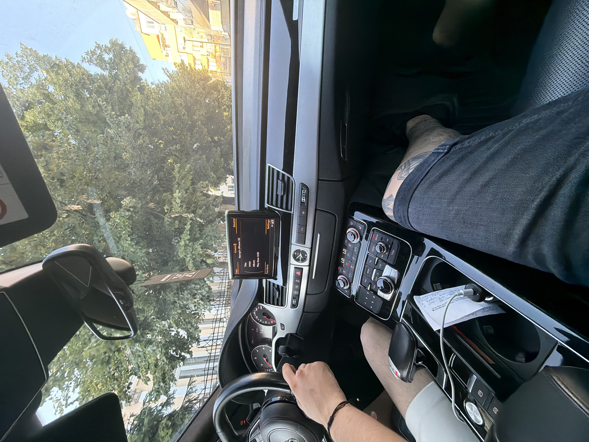 The image captures the interior of a car, likely an Audi, with two individuals inside. The driver, whose left hand is visible on the steering wheel, is wearing a white t-shirt and a black bracelet. Their right leg is also visible, partially covered by a white short. The passenger, on the right, is wearing dark jeans and has visible tattoos on their leg.
The car's dashboard is prominently displayed, featuring a central infotainment screen showing "George's iPhone (2)" as the media source, with song title, artist, and album information. Below the screen are climate controls, buttons, and a dial. A charging cable is plugged into a device in the center console, holding a piece of paper. The car appears to be moving, as indicated by the blurred trees and buildings visible through the windshield.
The exterior environment suggests a sunny day with a clear blue sky. Lush green trees line the street, and buildings with light-colored facades and orange rooftops are visible in the background. The architecture suggests an urban setting, likely in Ruse, Bulgaria, given the context provided. The overall scene conveys a sense of everyday travel.