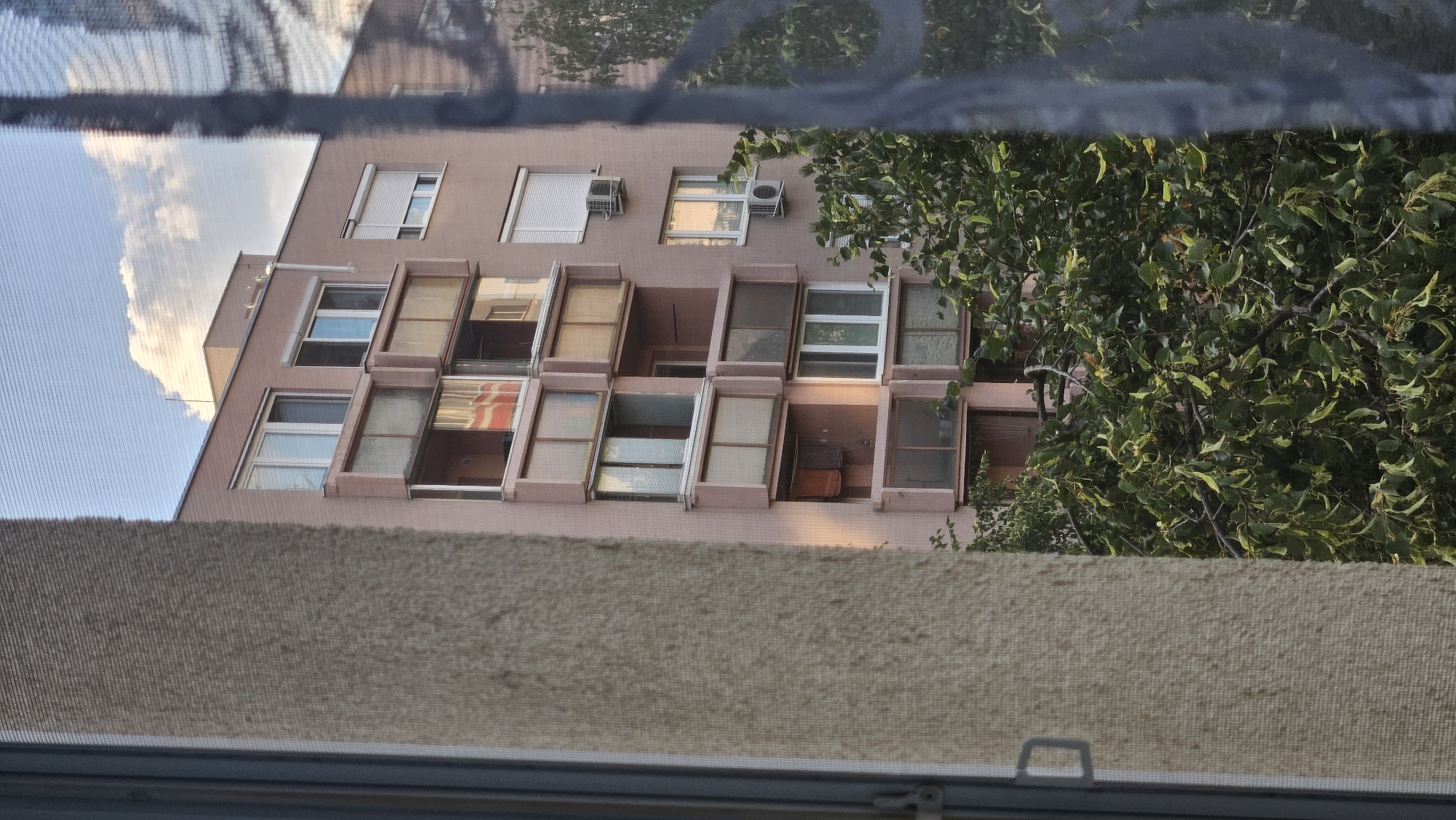 The image captures a view through a window with a screen, looking out at a pink apartment building in Bratislava, Slovakia. The building features multiple balconies with glass enclosures, some of which reflect the sky and clouds. There are several windows visible, some with blinds. Two air conditioning units are mounted on the exterior wall. Lush green trees partially obscure the lower floors of the building. The foreground shows a textured wall of the building or window frame and a dark curtain or screen. The sky is partly cloudy.