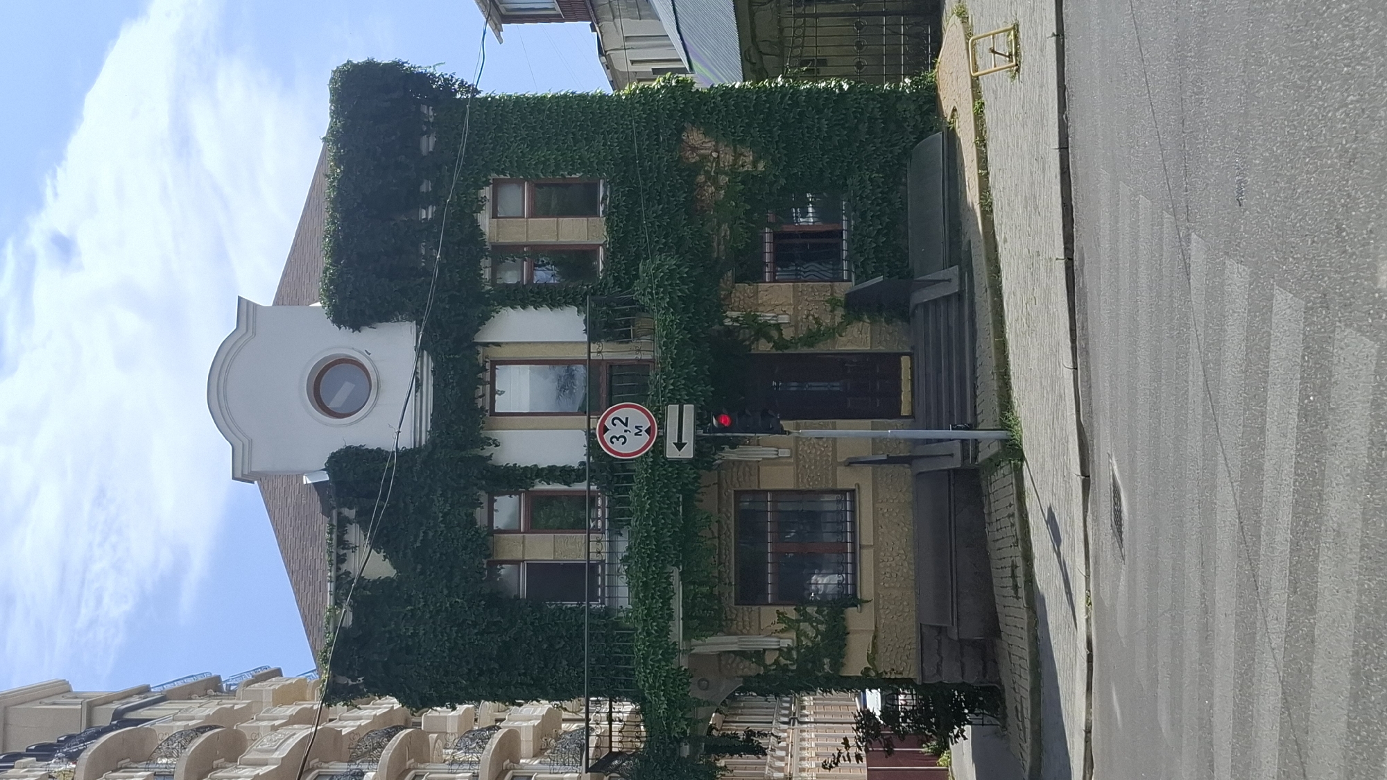 An outdoor, eye-level, wide shot captures a building covered in dark green ivy under a bright, partly cloudy sky. The building has a light-colored facade and a brown roof. A balcony with black railings spans the second story. There are at least three windows visible on the second story, with two on the first. A traffic light showing red hangs from a pole in front of the building, next to a circular sign with "3.2 M" inside and an arrow pointing left. A crosswalk is visible in the foreground on the asphalt road. To the left of the building, a portion of a taller, beige building with balconies is visible. To the right, a dark metal fence and the top of another building are seen.