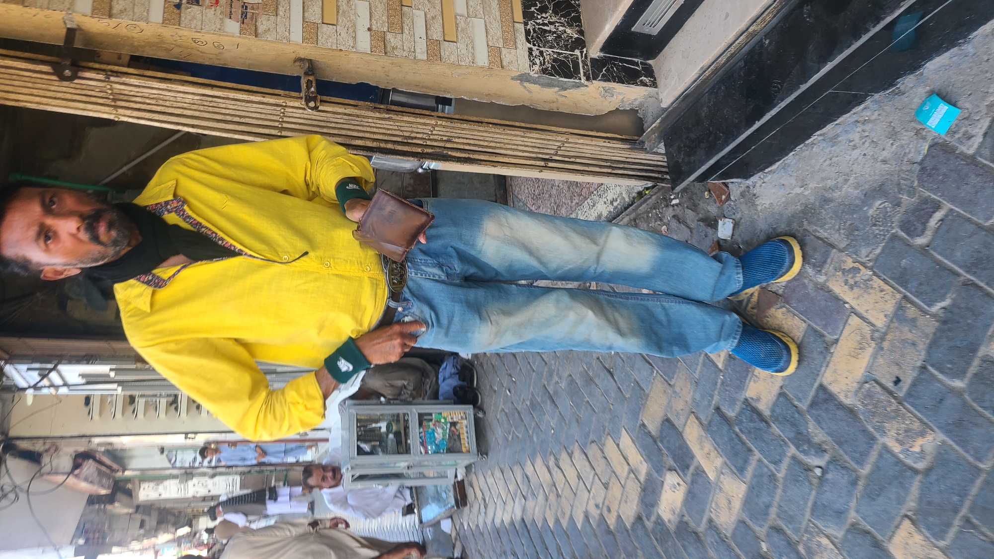 A man in a yellow shirt, blue jeans, and blue shoes with yellow soles stands on a brick-paved street. He is holding a brown wallet and looking toward the camera. Other people and vendor carts are visible in the background. The location is Quetta City Tehsil, Pakistan.