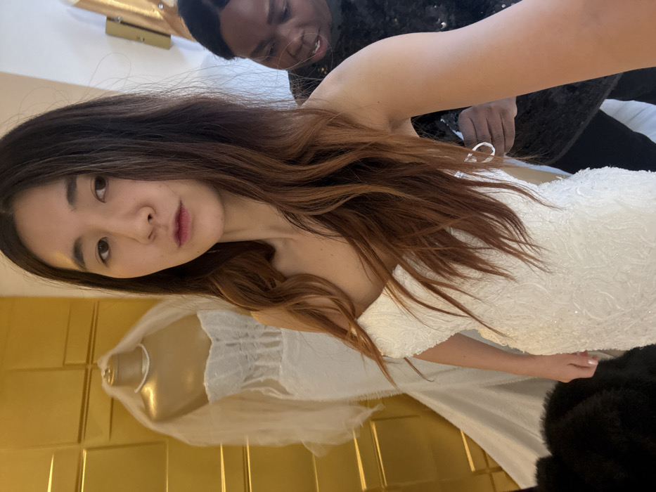 A woman with long brown hair wears a white, beaded dress. An African American person helps her with the dress. A mannequin wearing a wedding veil and a gold wall are also visible.