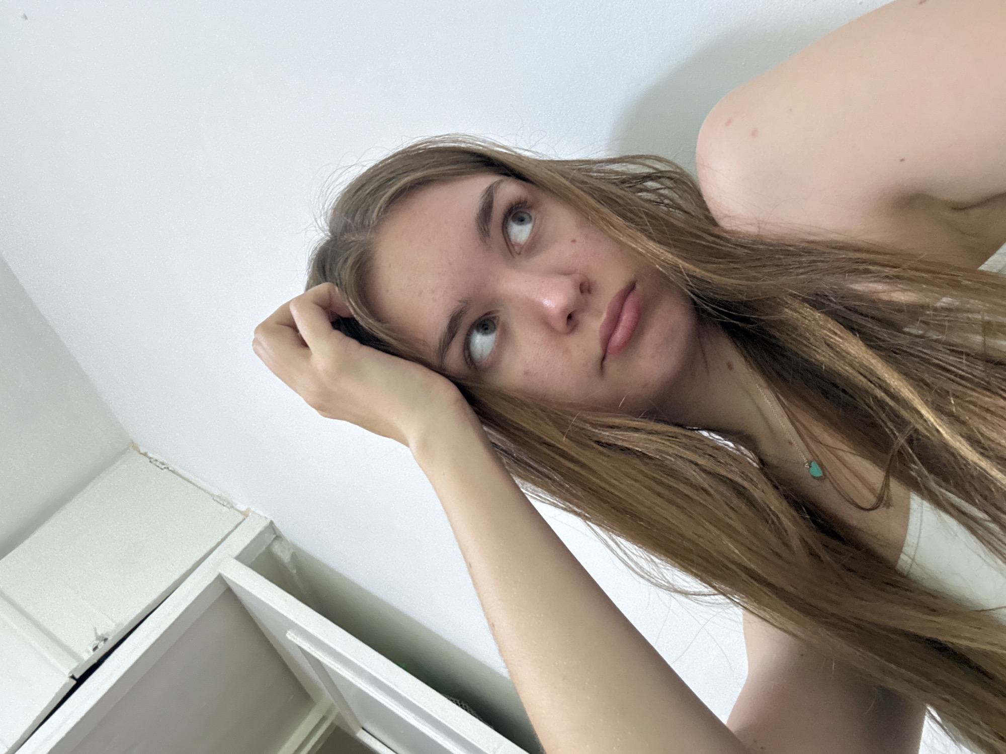 A fair-skinned young woman with long, light brown hair is lying down, propped up by her left arm. She is wearing a white tank top. Her gaze is directed upwards and to the left. A small turquoise pendant hangs from a necklace around her neck. A white cabinet is partially visible in the lower left corner of the image. The background is a plain white wall. The image does not contain enough information to determine a location in Perm, Russia.