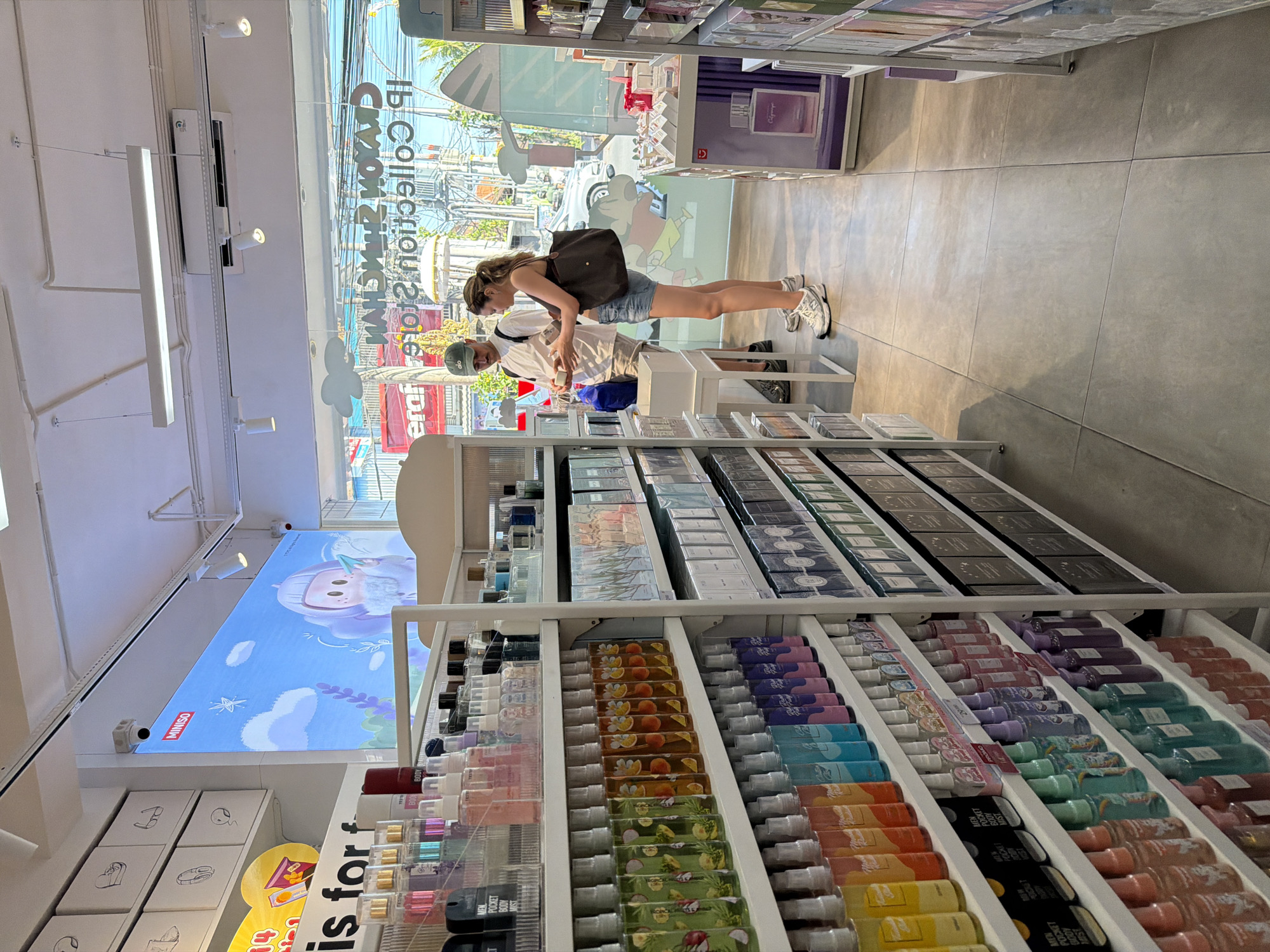 The image captures the interior of a retail store, likely a Miniso or similar brand, located in North Kuta, Indonesia. The store is well-lit with artificial lighting from overhead fixtures and a large digital screen displaying animated content. Shelving units line the aisles, displaying a variety of consumer products, primarily beauty and personal care items such as perfumes, body mists, and skincare.
In the foreground, a man wearing a white t-shirt, khaki shorts, and a green baseball cap stands next to a woman in a pink tank top and denim shorts, holding a tote bag. They appear to be examining a product together. The woman is wearing white sneakers, and the man is wearing dark athletic shoes. They are positioned between two shelving units filled with colorful spray bottles.
The store's exterior is visible through large glass windows, showing a street scene with vehicles and buildings, indicating an urban or semi-urban setting. The flooring is tiled in a light gray color. The overall impression is that of a bright, modern retail environment.