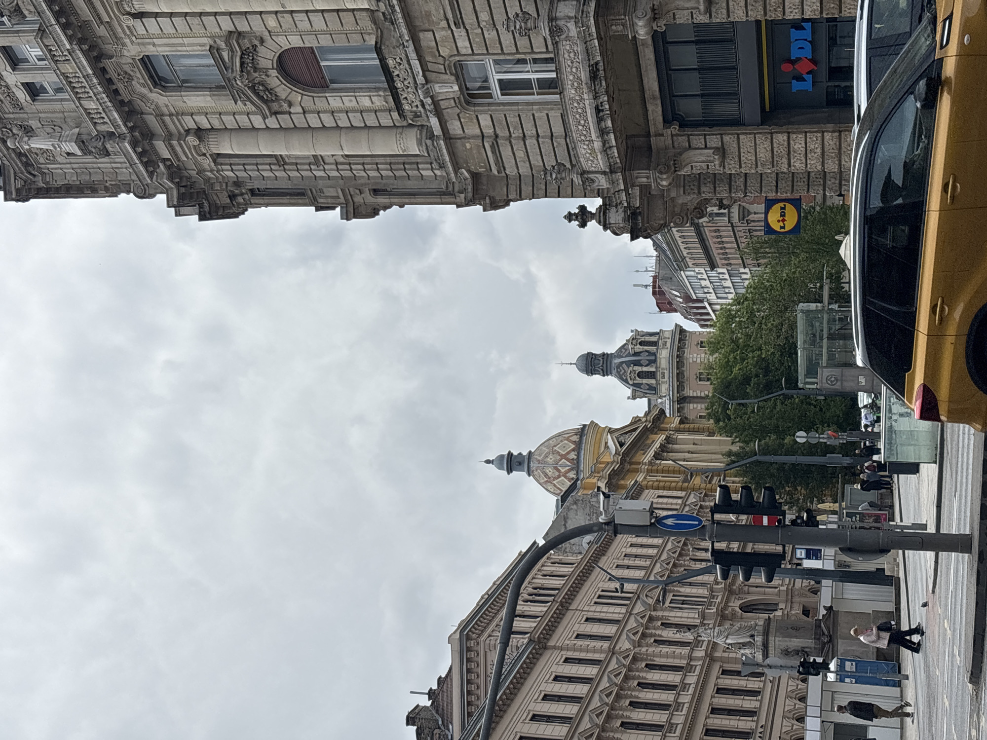 The image depicts a street scene in Budapest, Hungary, characterized by ornate, historic buildings lining the thoroughfare. The sky is overcast.
On the right side of the image, a yellow car is partially visible. Street furniture, including traffic lights and signs, is present. People are walking on the sidewalk. A Lidl supermarket sign is seen.
In the background, a building with a notable dome is visible amidst trees and other structures.
The image conveys an urban atmosphere with a blend of architectural heritage and modern city life.