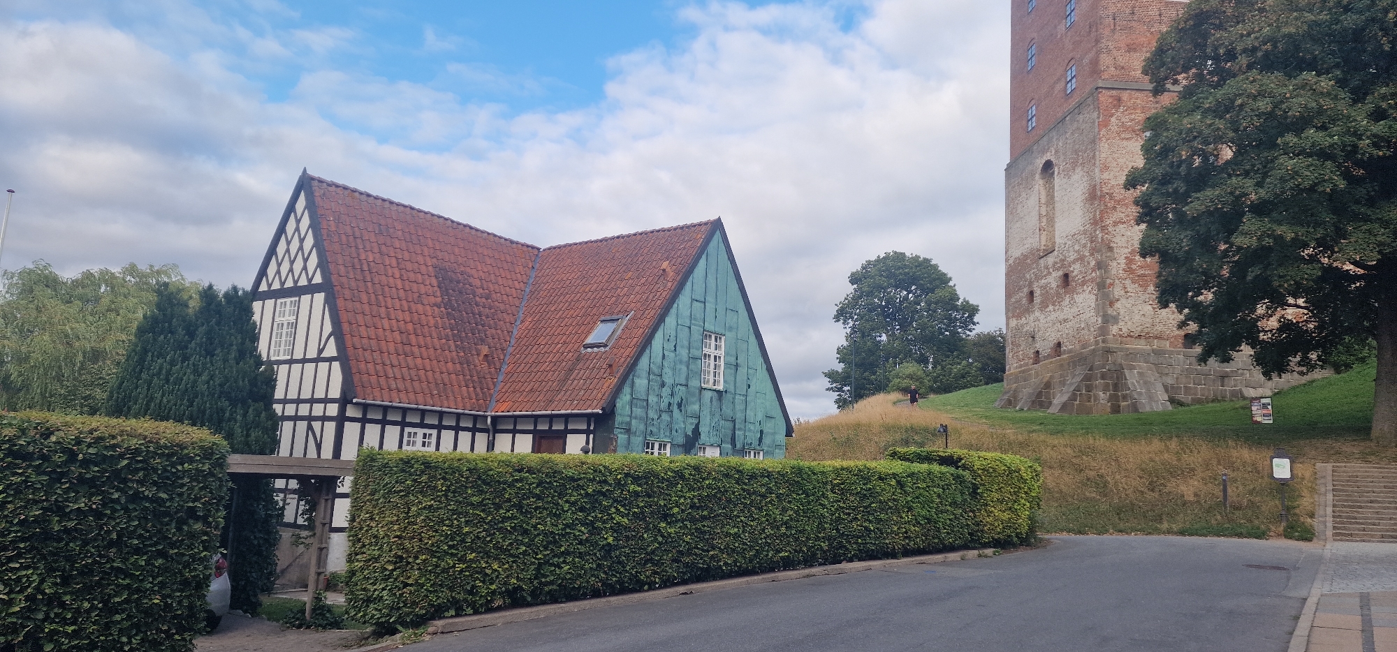 The image depicts the Koldinghus castle tower in Kolding, Denmark, viewed from a lower vantage point. To the left of the tower is a traditional Danish half-timbered house with a red tiled roof. In front of the house, a neatly trimmed hedge runs horizontally across the frame, obscuring the lower portion of the house and a driveway. A car is partially visible behind the hedge. The ground leading up to the castle tower is covered in dry, brown grass, with a few trees and patches of green lawn. A set of stone stairs is visible to the far right, leading up the embankment. The sky is partly cloudy with blue sky showing through. Two individuals are visible on the grassy hill: one standing and the other appearing to be a dog.