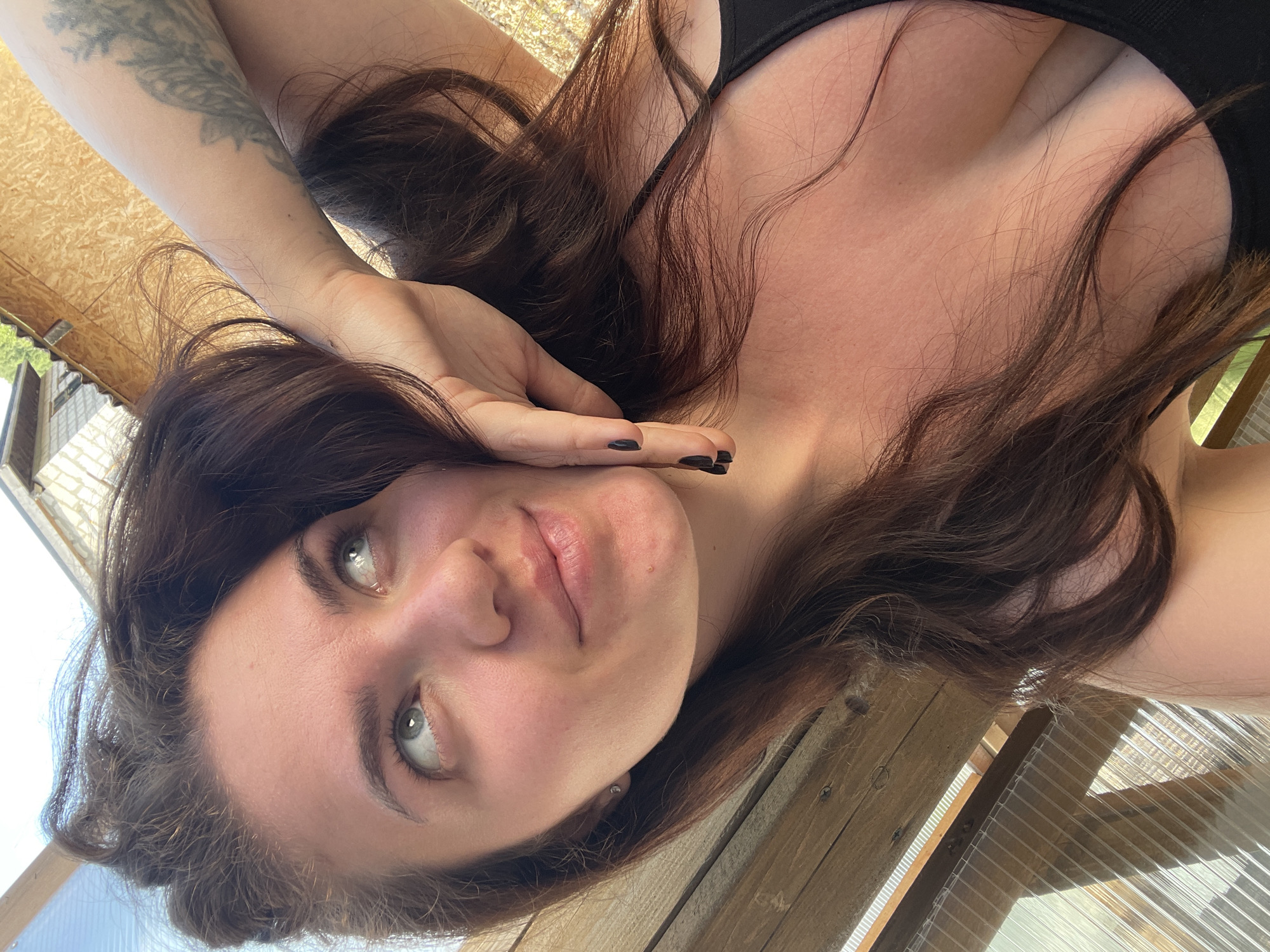 A woman with light skin and long brown hair is lying down, looking up at the camera. She is wearing a black top with thin straps. Her left arm, which has a tattoo, is raised with her hand near her face, displaying dark nail polish. The background appears to be a partially constructed or rustic wooden structure, possibly within a greenhouse or similar outdoor setting. The location is not discernible from the image, and there is no evidence to suggest it is in Мосметаллоконструкция Онт, Russia.