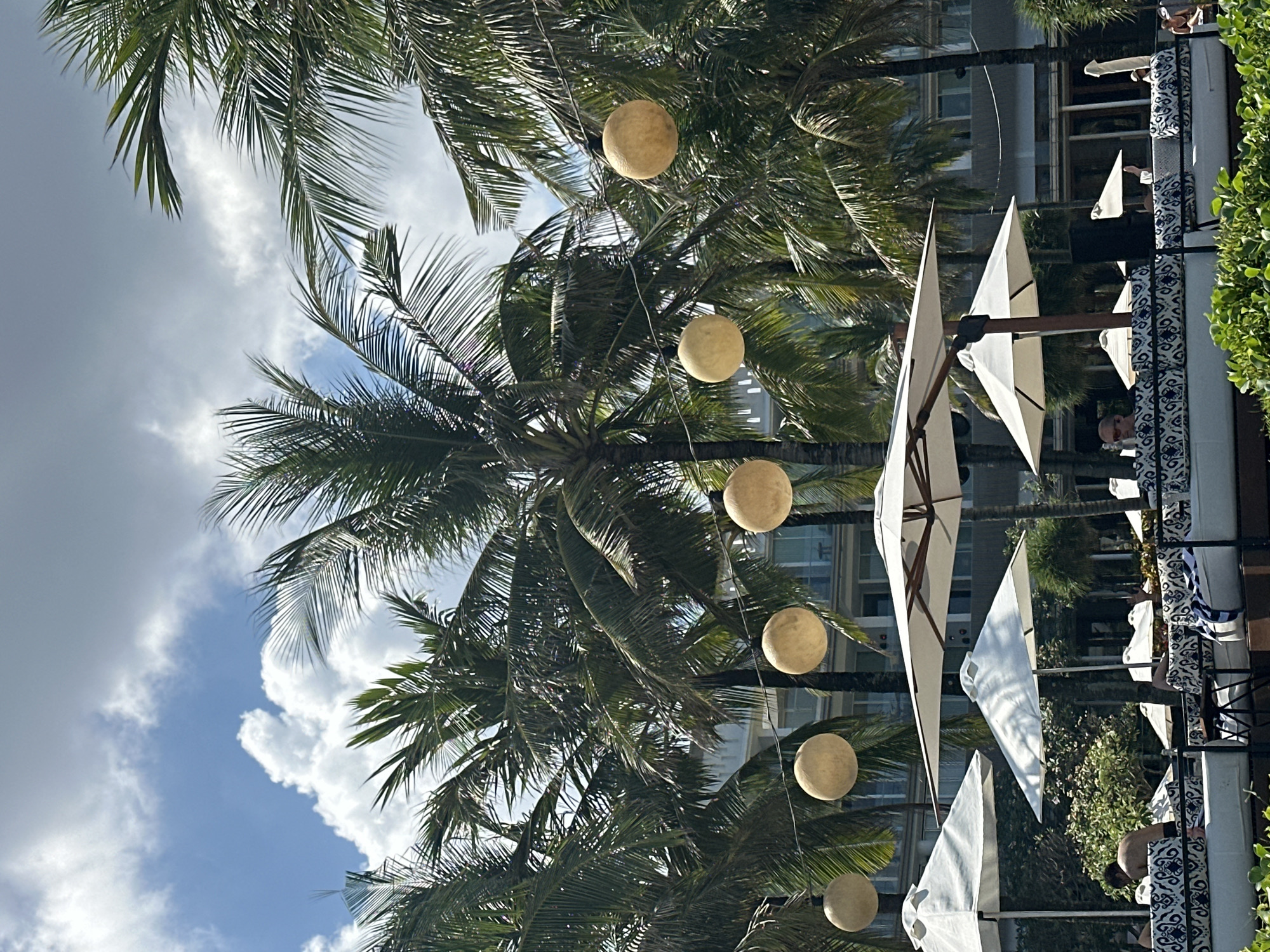 The image captures a scene at a resort or outdoor lounge area in North Kuta, Indonesia, on a bright, sunny day. The foreground is dominated by lush green foliage, hinting at a tropical setting. Behind this, several white lounge beds with patterned blue and white cushions are arranged. A few people are visible reclining on these beds, some partially obscured.
Overhead, a string of large, spherical lights, appearing to be lanterns, hangs suspended between tall palm trees. These palm trees are abundant throughout the scene, their fronds rustling gently, framing the view of a modern building in the background. The building appears to be a hotel or resort, with multiple floors and balconies.
Several white umbrellas are open, providing shade over some of the seating areas. The sky above is a vibrant blue, dotted with fluffy white clouds, indicative of fair weather. The overall impression is one of a relaxed, tropical vacation destination.