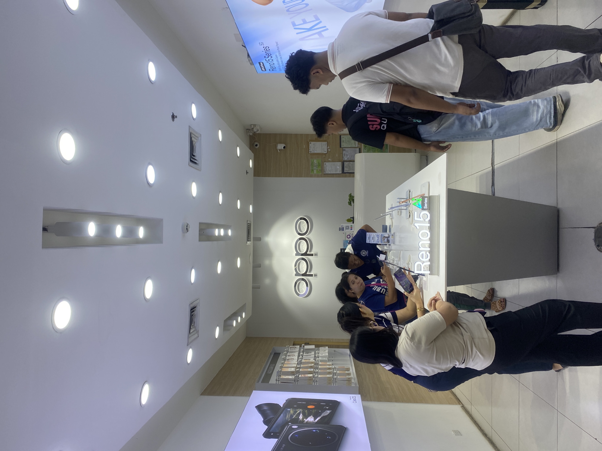The image depicts the interior of an OPPO electronics store, likely located in Davao City, Philippines. Several individuals, a mix of customers and possibly staff, are gathered around display tables showcasing mobile phones. A large digital screen on the left displays an advertisement for an OPPO product, featuring a camera. Another screen on the right shows an advertisement for the "Reno 15 Series 5G". The store is well-lit with overhead fluorescent lights and has a clean, modern aesthetic with white walls and display counters. The floor is tiled with a light-colored pattern. The overall scene captures a typical retail environment for consumer electronics.