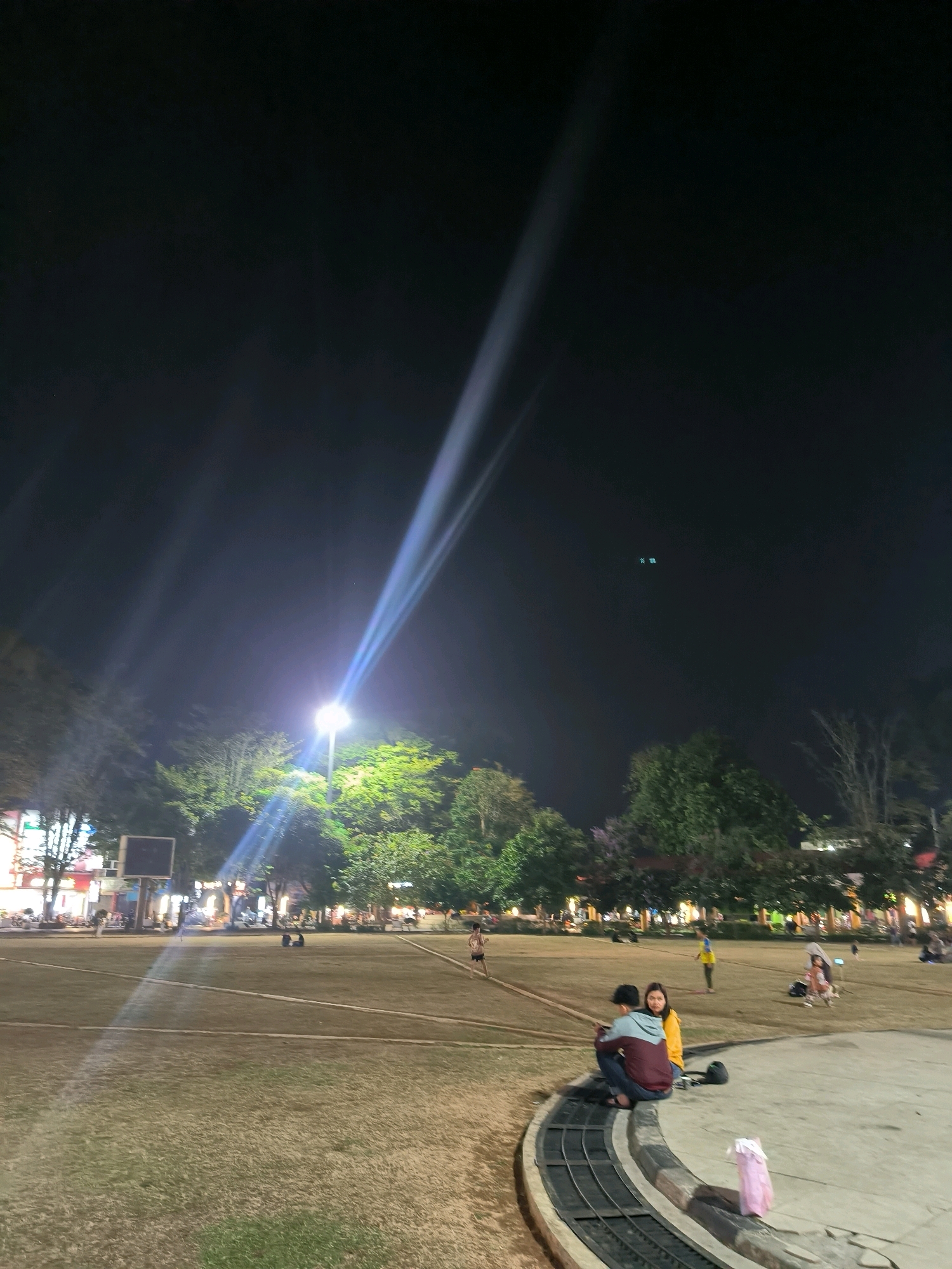 This image captures a nighttime scene at a park in Salatiga, Indonesia. A bright lamppost illuminates the sky with a strong beam of light, casting long shadows across the grassy field. Several people are scattered throughout the park, some sitting on a curved concrete structure in the foreground, while others are walking or standing in the background. Trees and buildings are visible behind the park, suggesting an urban setting. The overall atmosphere is one of casual evening activity in a public space.