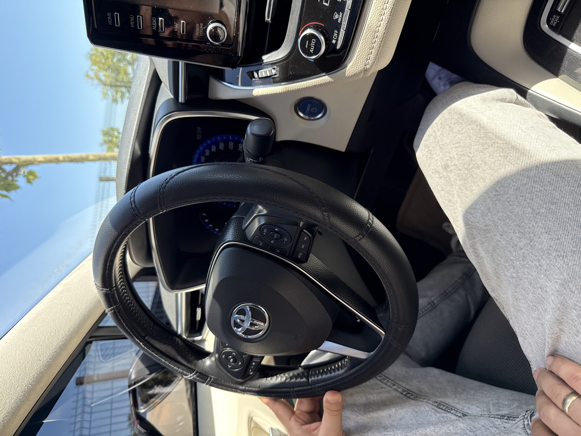 The image shows the interior of a car, with a person's hands and legs visible. The focus is on the steering wheel and the dashboard. The steering wheel is black leather with a Toyota logo in the center. There are buttons on the steering wheel for controlling audio and other functions. The dashboard features a digital display showing the time as 10:59 and a speedometer. To the right of the steering wheel is a touchscreen infotainment system with options for HOME, MENU, AUDIO, and MAP. Below the screen are climate control buttons and a POWER VOLUME knob. A blue START/STOP button is visible on the center console. The interior appears to be beige and black. The person is wearing light-wash jeans. The scene is lit by sunlight, suggesting it is daytime. The background shows a blurry view of trees and a fence through the car window, consistent with an outdoor setting in a place like Sancaktepe, Turkey.