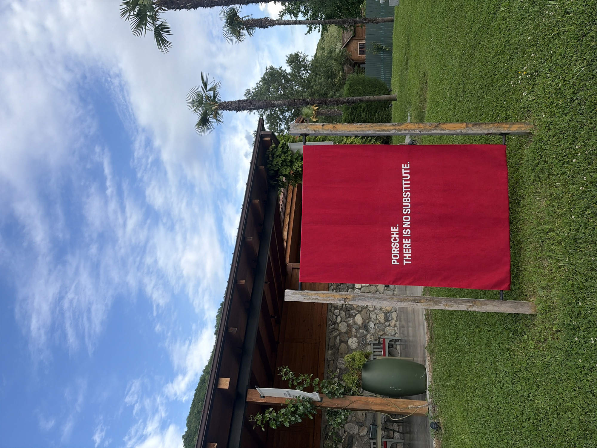 The image captures a scene in Napareuli, Georgia, under a partly cloudy sky. A large, rectangular red banner is prominently displayed, bearing the text "PORSCHE. THERE IS NO SUBSTITUTE." in white capital letters. The banner is attached to two wooden posts, set on a green lawn.
Behind the banner, there's a building structure, possibly a part of a house or a canopy, with a stone-covered wall. A green, elongated container is also visible.
Palm trees stand tall in the background, adding a contrasting element to the Georgian landscape. Further back, there's a wooden house and green foliage, suggesting a rural or suburban setting. The environment includes grass, sky, and various architectural and natural elements, all characteristic of the Napareuli region.