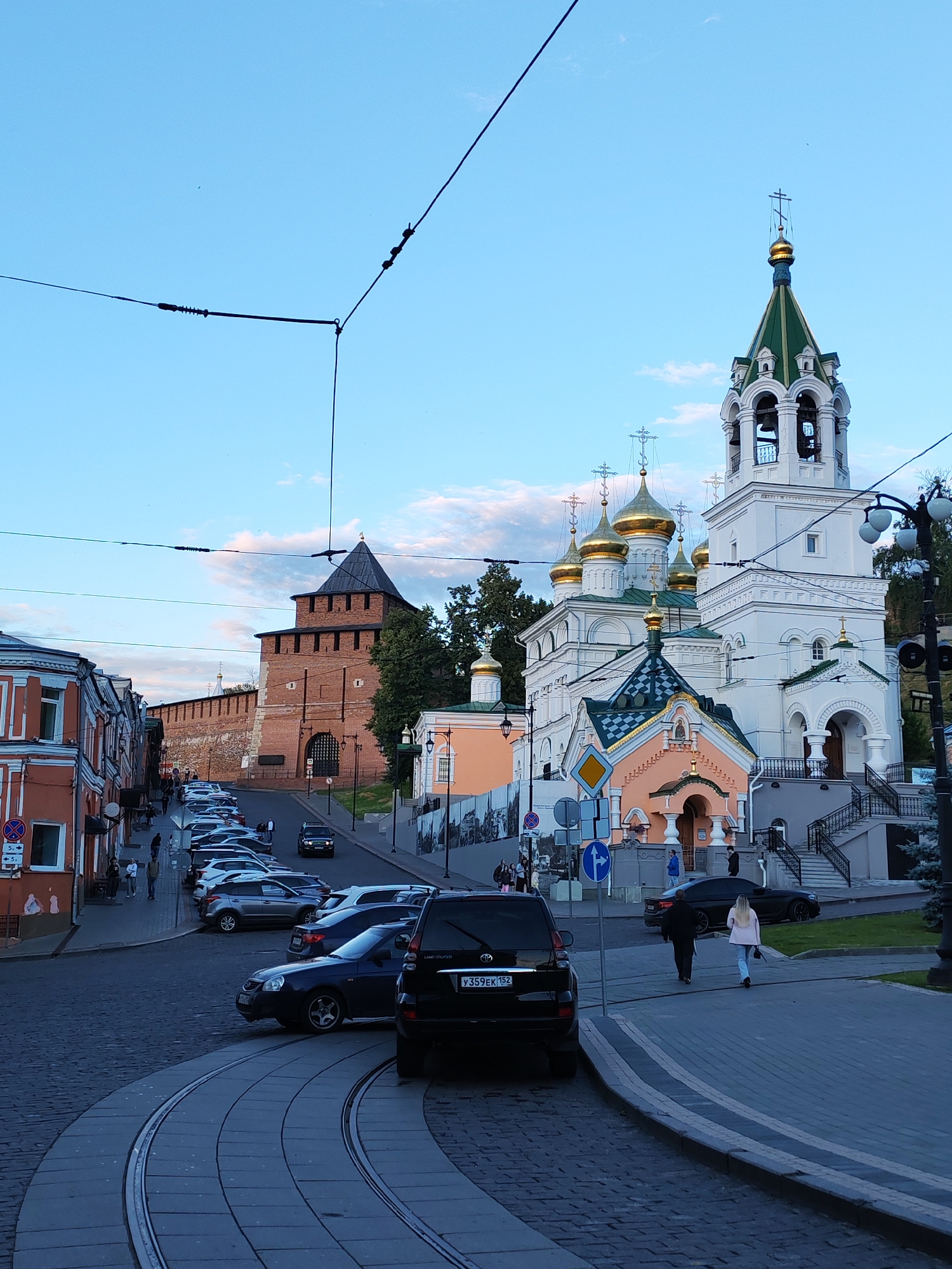 The image captures a street scene in Nizhny Novgorod, Russia, featuring a mix of historical and modern elements under a twilight sky. Prominently in the foreground are tram tracks curving along a cobblestone street, with several parked cars lining the right side of the road.
Further down the street, a large brick fortress wall with a prominent tower rises up, suggesting historical significance. Adjacent to the wall is a vibrant orange building with multiple windows. To the right, a white church with multiple golden domes and a green-topped bell tower stands majestically, adorned with crosses. A smaller, pastel-colored structure with a patterned roof is situated in front of the main church.
Several people are visible walking on the sidewalks and near the parked cars, indicating a calm, everyday activity. The sky is a soft blue with hints of pink and white clouds, suggesting the late afternoon or early evening. The lighting is soft, casting gentle shadows and creating a peaceful atmosphere.
Visible text includes license plates on the cars, such as "У359ЕК 152," and traffic signs with Russian lettering and symbols. The overall scene conveys a sense of place, blending the ancient architecture of the Kremlin walls with the active life of a modern city.
