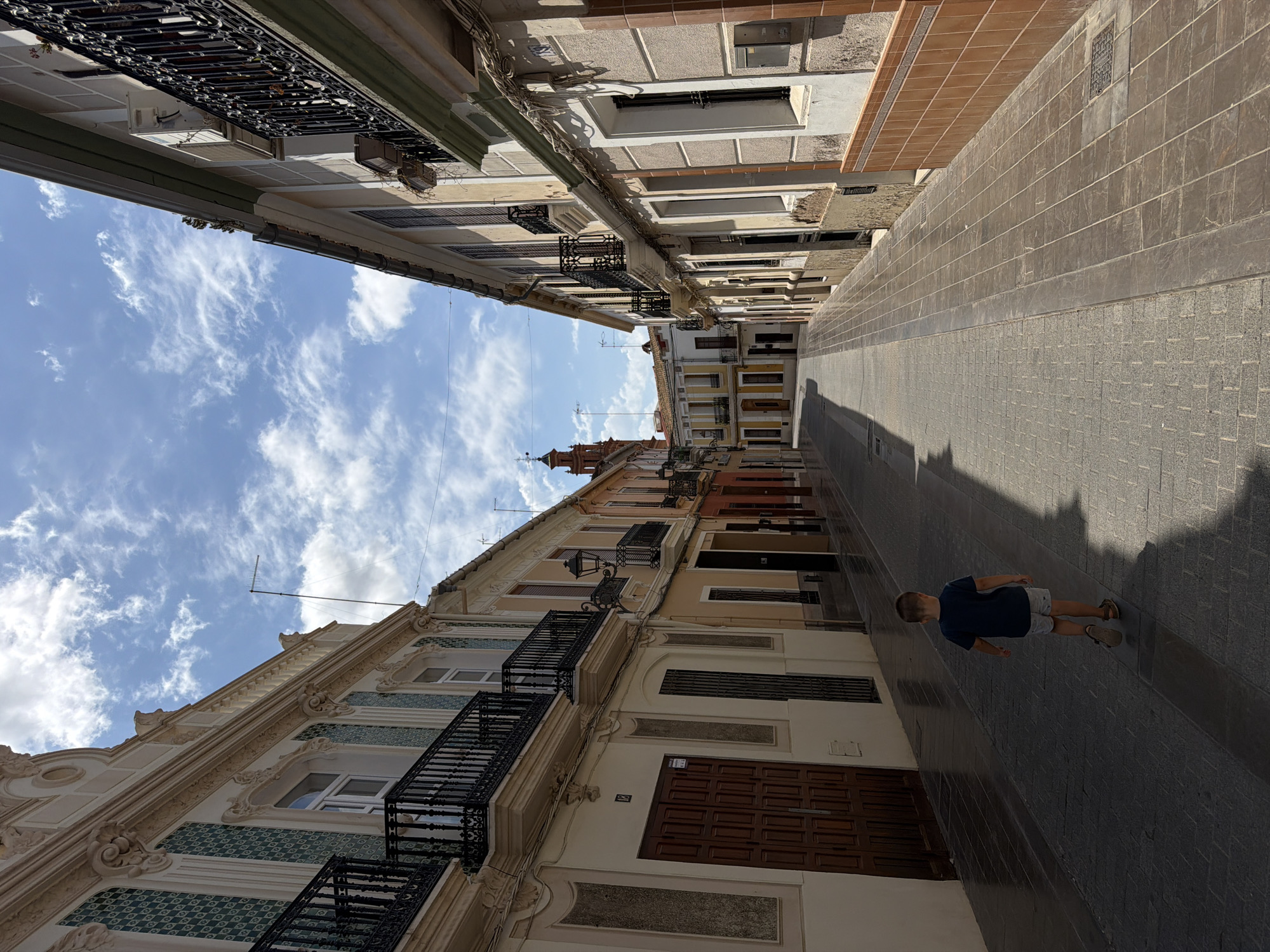 A lone child walks down a narrow street lined with historic buildings. The child, a young boy wearing a dark blue t-shirt and light-colored shorts, is walking away from the camera towards the vanishing point of the street. The street is paved with cobblestones and tiles, with a shaded area on the left side and a sunlit area on the right. The buildings on either side of the street are multi-story structures with ornate balconies, shutters, and tiled facades. The sky is blue with scattered white clouds. In the distance, a church tower is visible. The overall impression is of a quiet, sunny afternoon in a European city.