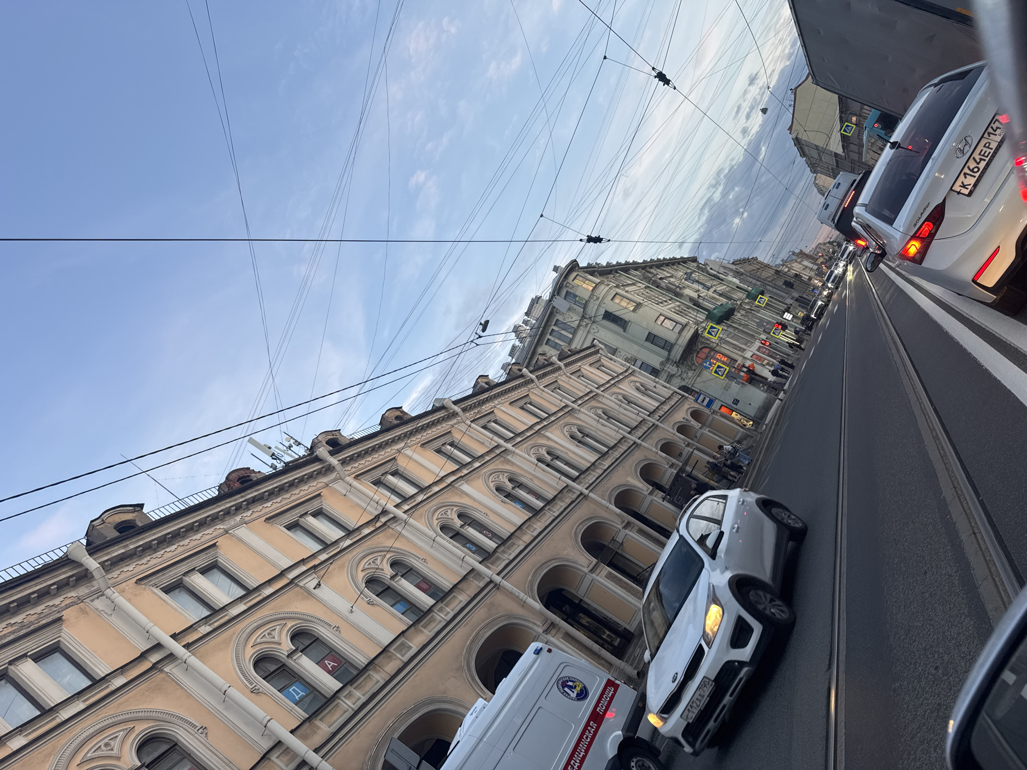 This image captures a tilted view of a busy street in Saint Petersburg, Russia, showing urban architecture and vehicle traffic under a partially cloudy blue sky.
In the foreground, a white Hyundai Solaris with Russian license plate K 164 EP 147 RUS is visible from the rear, its brake lights illuminated, suggesting it is stopped or moving slowly in traffic. To its left, a white Kia Ceed with license plate E 112 TA 750 RUS is seen from the front-side, also in motion. Directly in front of the Kia, a white van displaying "Скорая медицинская помощь" (Emergency Medical Service) and "03" with a medical logo is partially visible. Tram tracks run along the road, indicating a route for public transport.
The street is lined with grand, multi-story buildings, characteristic of Saint Petersburg's historical architecture. One prominent building on the left is light beige/yellow, featuring numerous arched windows, intricate cornices, and decorative elements. Visible within some of its windows are Cyrillic letters like "A", "д", "м", and "Э", possibly from signs or displays inside. Another building further down the street on the right is lighter in color, possibly light grey or blue. Overhead, a complex network of power lines, likely for trams or trolleybuses, crisscrosses the sky.
The scene depicts a daytime setting with sufficient natural light. Traffic lights and pedestrian crossing signs are visible along the street. A few pedestrians can be seen in the distance near the crossings. The overall impression is one of a typical urban street scene in motion.
