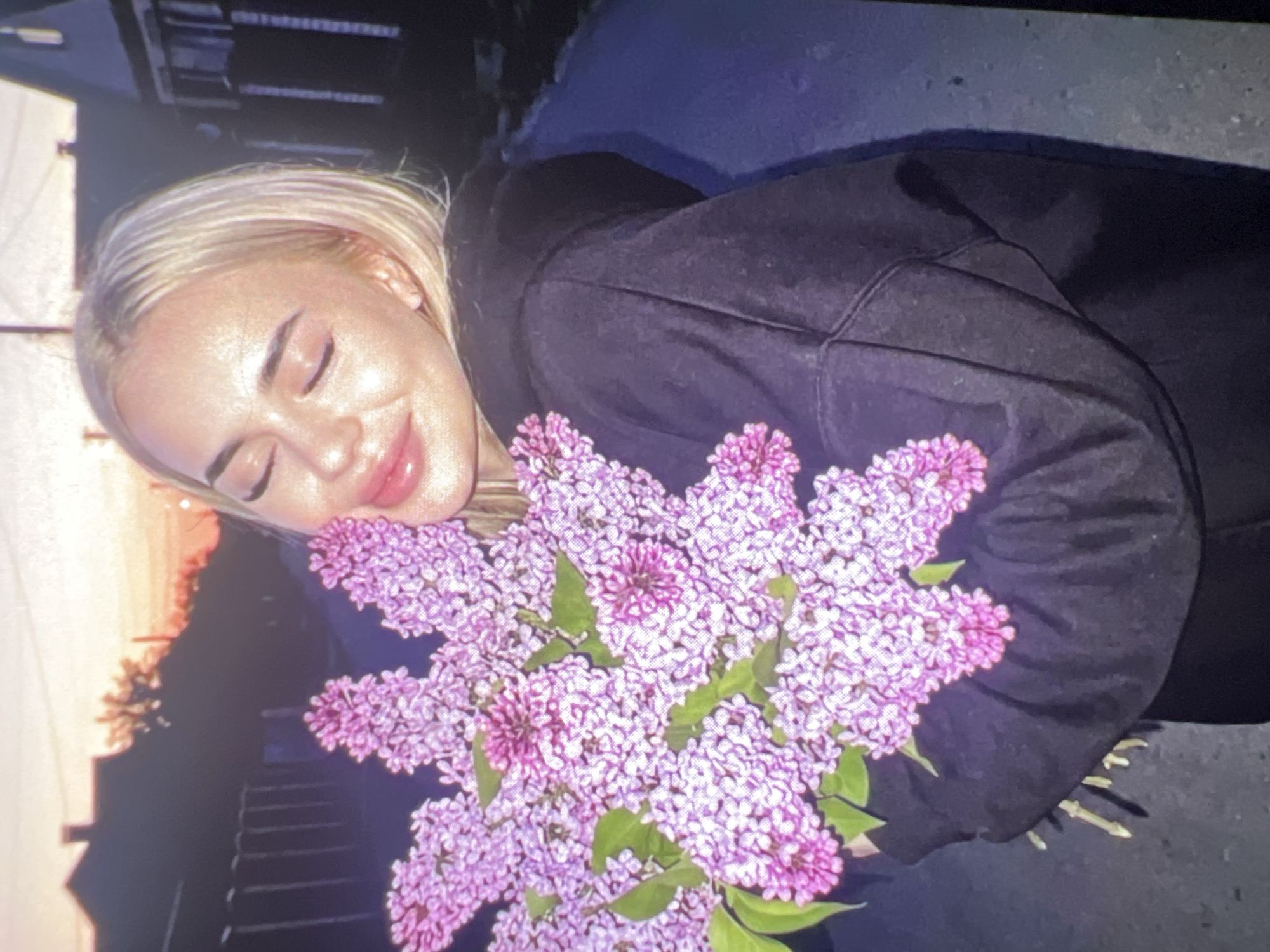 A young woman with blonde hair and light skin is depicted in a portrait-style shot, leaning her head onto a large, vibrant bouquet of lilac flowers. Her eyes are closed, and a gentle smile plays on her lips, conveying a sense of serenity and happiness. She is wearing a dark, possibly black, hoodie or jacket, which contrasts with her brightly lit face and the colorful flowers.
The bouquet consists of numerous clusters of purple and pink lilac blooms, dense and full, with some green leaves visible among them. The flowers are held close to her upper body, obscuring part of her clothing.
The background is softly blurred but suggests an outdoor setting during either sunrise or sunset, indicated by a warm, orange-pink glow on the left side of the frame, transitioning to darker tones. Subtle outlines of what appear to be buildings or structures are visible in the distance. The lighting is soft and warm, highlighting the woman's face and the flowers, giving them a radiant quality. The overall image appears to be a photograph taken of a digital screen, as evidenced by visible scan lines or pixel patterns throughout the picture. There is no discernible text in the image.