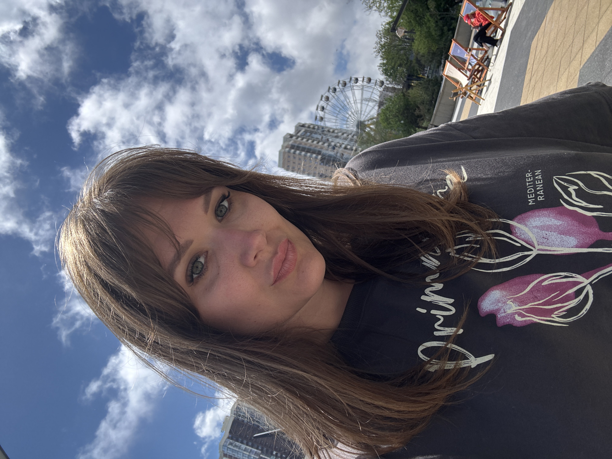 A woman is facing the camera for a selfie. She has long, light brown hair with bangs, green eyes, and fair skin. She is wearing a black sweatshirt with a floral design. In the background, there are buildings, trees, a Ferris wheel, blue sky with clouds, and lounge chairs on a deck. The location is Novorossiysk, Russia.