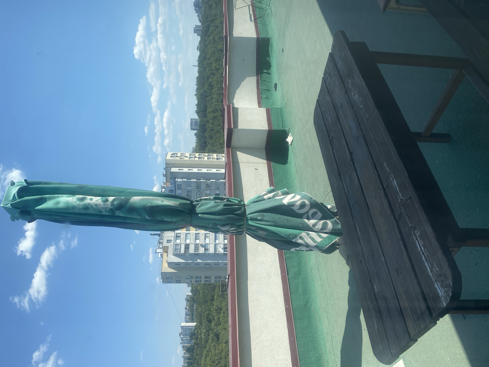 The image shows a closed green umbrella on a rooftop terrace in Chișinău, Moldova. The umbrella is patterned with white circular shapes and text that is difficult to read. A dark wooden picnic table is in the foreground, casting a shadow on the green-covered terrace. In the background, there are several multi-story buildings and a dense line of trees, indicating an urban environment. The sky is bright blue with scattered white clouds. The overall scene depicts an outdoor seating area on a sunny day.