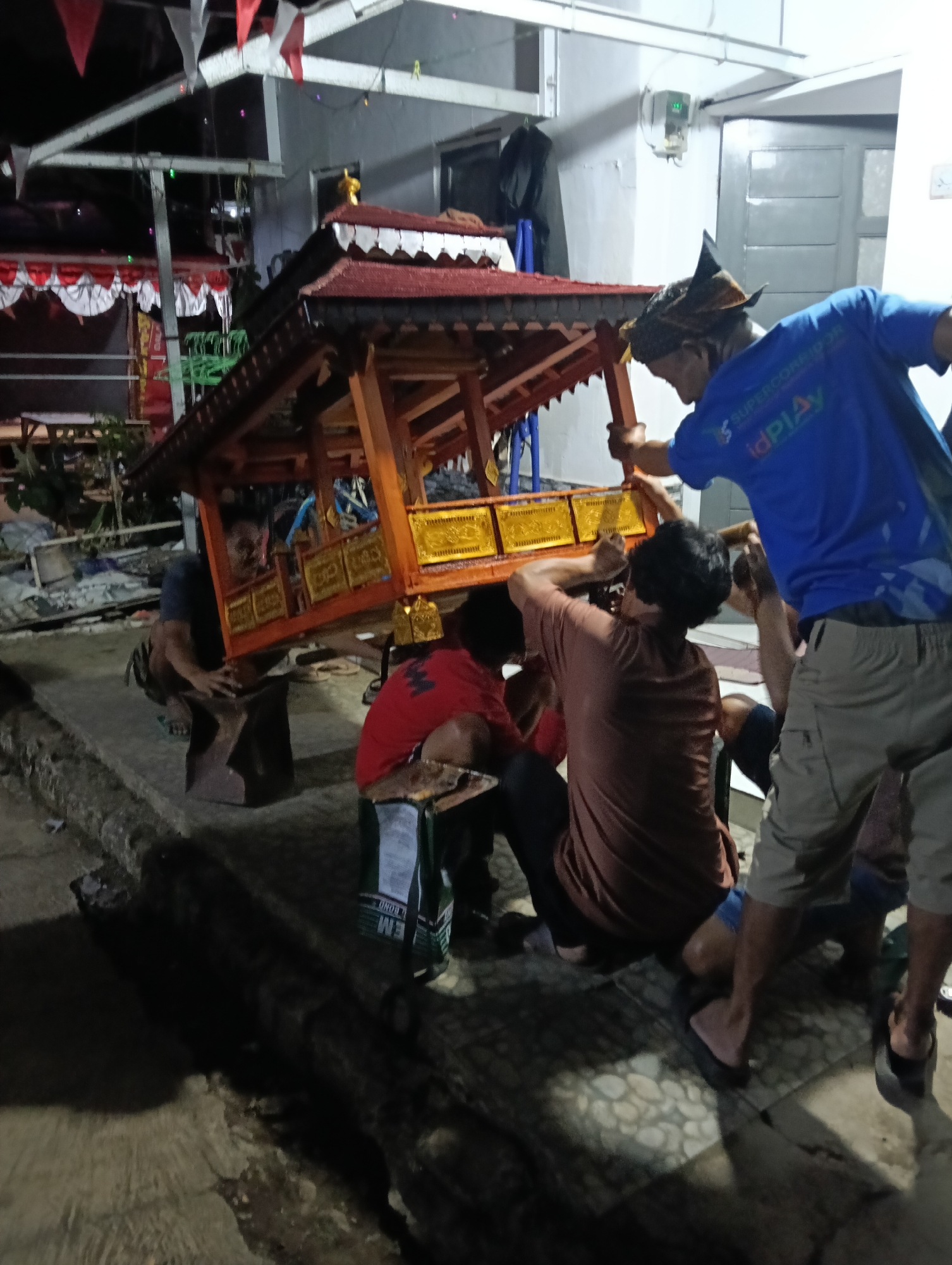 The image depicts a group of men working together to move a wooden structure, possibly a decorative float or altar, outdoors at night. The structure has red and gold ornamentation and appears to be quite heavy, as multiple individuals are actively supporting and maneuvering it. One man, wearing a blue shirt with a logo and a traditional headdress, is seen from the side, holding onto the structure. Another man in a brown shirt is directly under it, supporting its weight. A third person in a red shirt is also involved in the effort. The environment appears to be a paved area, possibly a street or courtyard, with some foliage and buildings visible in the background, including what looks like a house with a closed door. Flags and bunting are hung in the upper left portion of the image, suggesting a celebratory or ceremonial event. The ground is concrete, and there are some discarded items, including a green bag, on the ground. The lighting suggests artificial illumination, creating shadows. The overall impression is one of communal effort in an outdoor, possibly informal, setting within Tasikmalaya, Indonesia.
