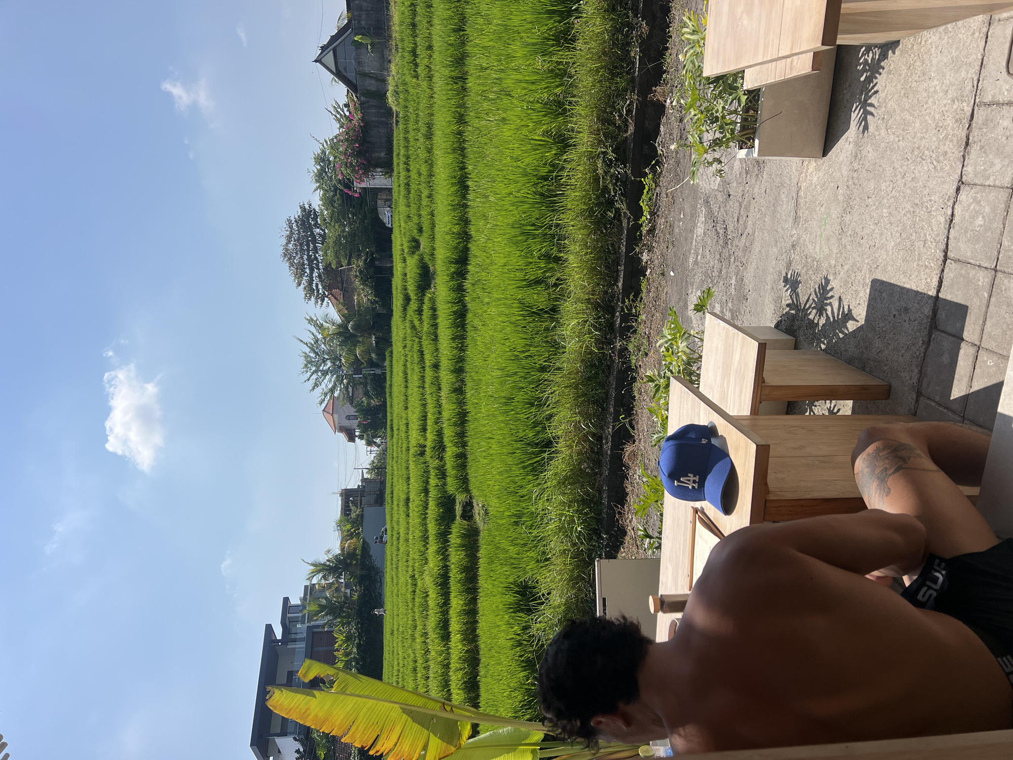 The image depicts an outdoor seating area with a view of lush green rice fields in Mengwi, Indonesia. In the foreground, a person is seated at a wooden table, facing away from the camera, with their back and tattooed leg visible. On the table, there is a blue baseball cap with a white logo, a menu, and a bottle. Two wooden benches are also present. Beyond the seating area, terraced rice paddies stretch out under a bright blue sky with scattered clouds. Several modern buildings and houses are visible in the background, interspersed with trees and vegetation. The overall scene suggests a serene and picturesque setting in a tropical environment.