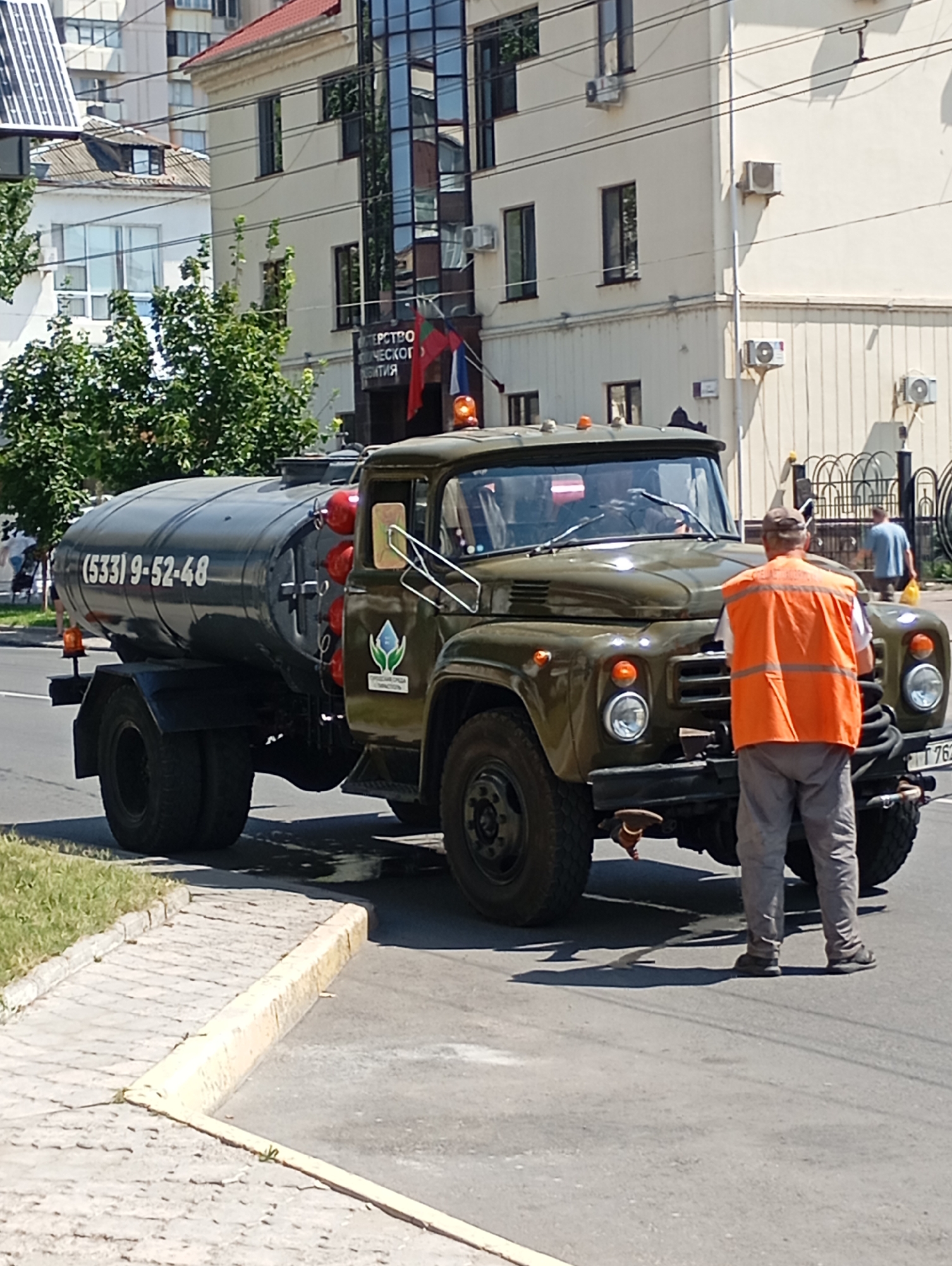 In Tiraspol, Moldova, on a sunny day, a green cistern truck with the phone number (533) 9-52-48 printed on its side is parked on a paved road. A person wearing an orange safety vest stands in front of the truck, facing it. Behind the truck, a building with architectural features typical of municipal buildings in Tiraspol, including multiple windows and air conditioning units, is visible. The building has flags flying outside. The scene is observed from a slight downward angle, capturing the truck, the person, and the urban environment.