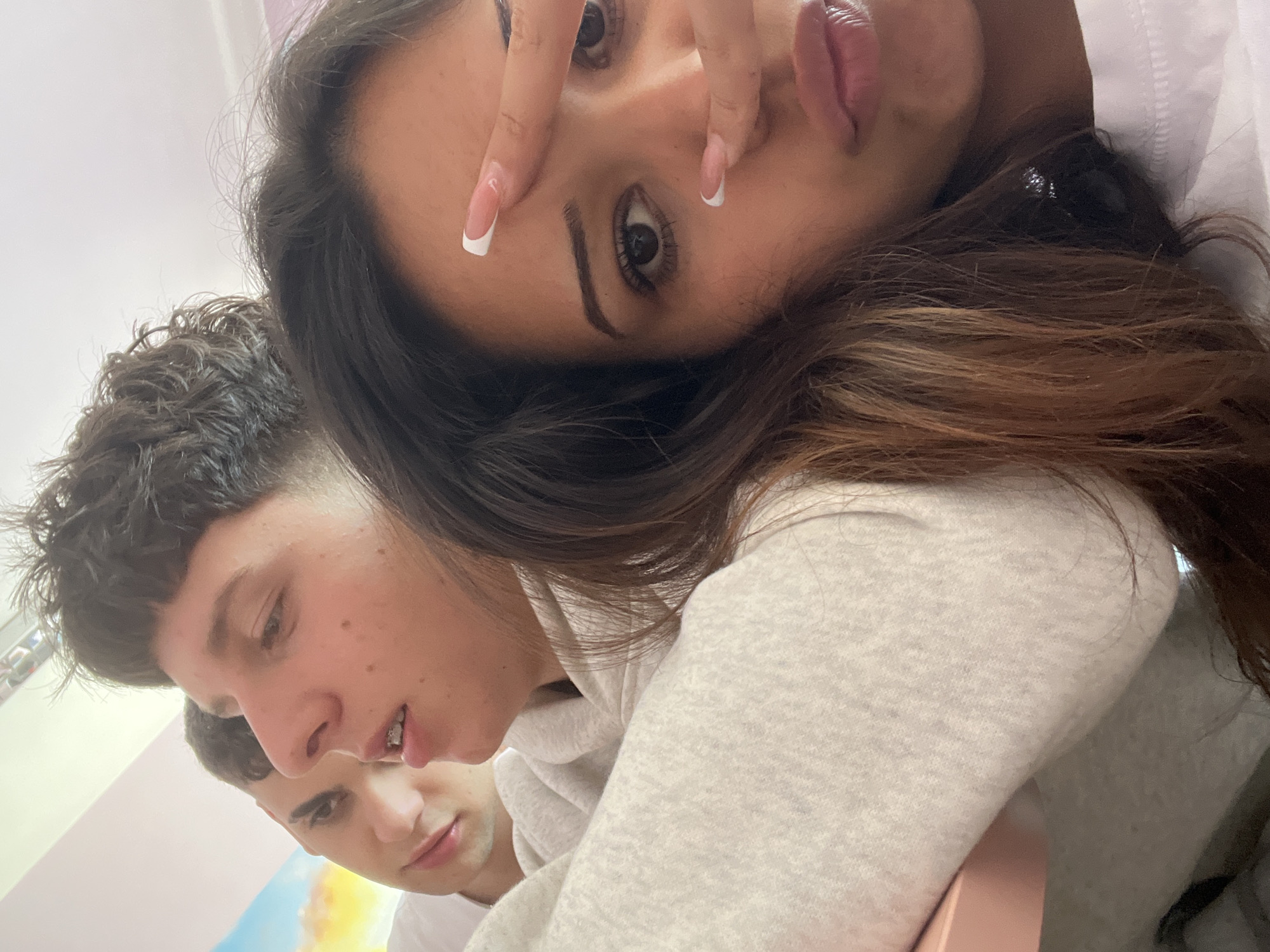 A casual, close-up indoor shot features three young individuals. The primary subject, a female, occupies the foreground, her face partially angled towards the camera. Her dark hair cascades around her, and she wears a light-colored, possibly grey, hooded top. She makes a playful 'peace sign' gesture with her fingers over her right eye, showcasing long nails with white French tips. Her lips are full and pink.
Behind and to her left, a male with dark, short, curly hair is visible, looking downwards with a slightly open mouth. He has small freckles or moles on his face and appears to be wearing a light-colored top. Further back and below him, only the face and shoulder of a second male are discernible, looking straight ahead with dark hair and a white t-shirt. The background appears to be a light-colored wall, transitioning from white to a light pinkish-blue and then to a yellowish-orange hue in the lower left, possibly a painted wall or abstract artwork. The image is rotated, giving it a dynamic, candid feel, with bright, even lighting throughout. No text is visible in the image.
