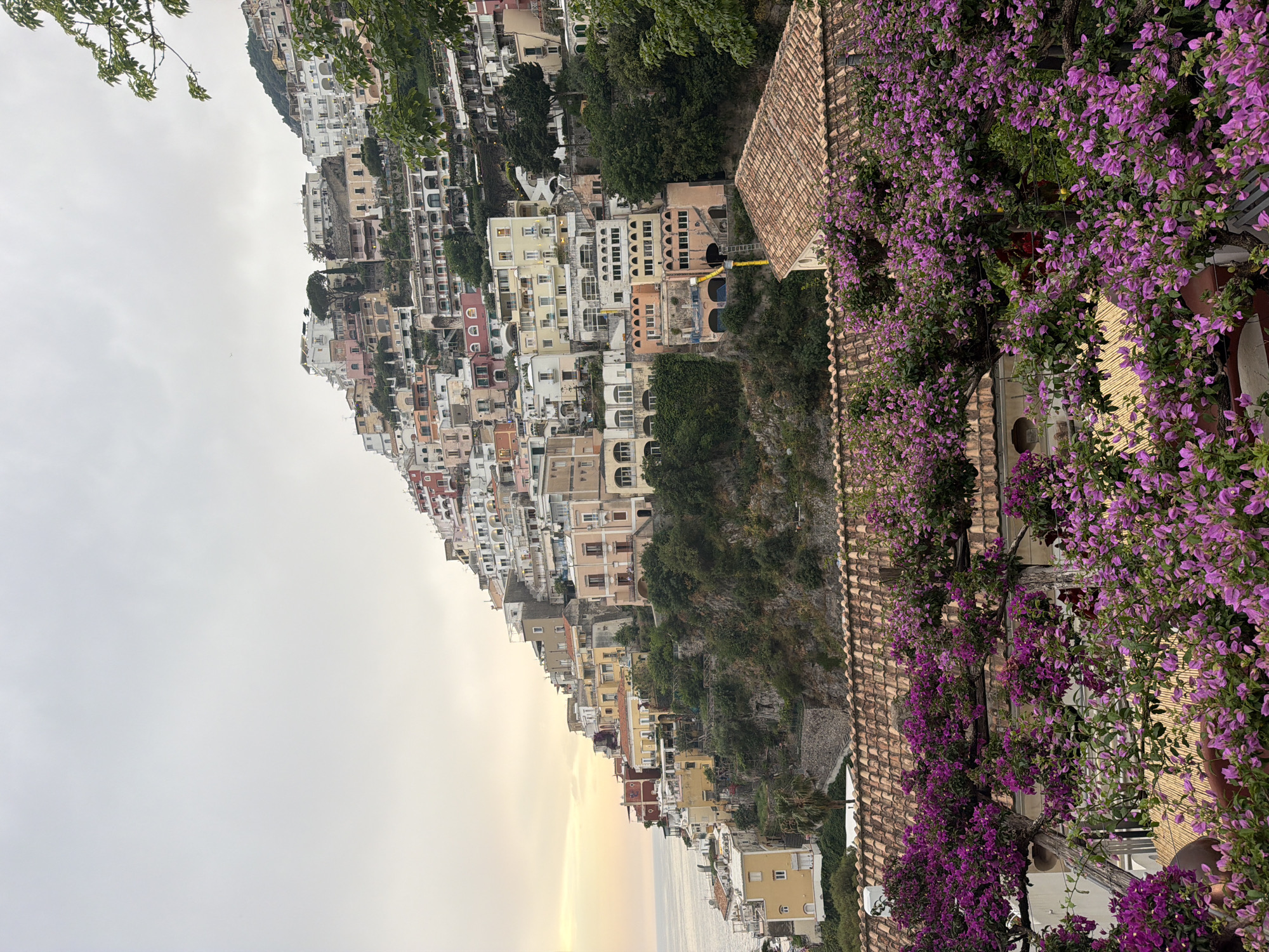 The image shows a view of Positano, Italy. The town is built on a steep hillside, with buildings stacked atop each other. The buildings are painted in various colors, including yellow, orange, pink, and white. The sky is overcast, with a hint of yellow near the horizon. To the right of the image, there is a building with a tile roof covered in purple bougainvillea flowers. There are no people visible in the image.
