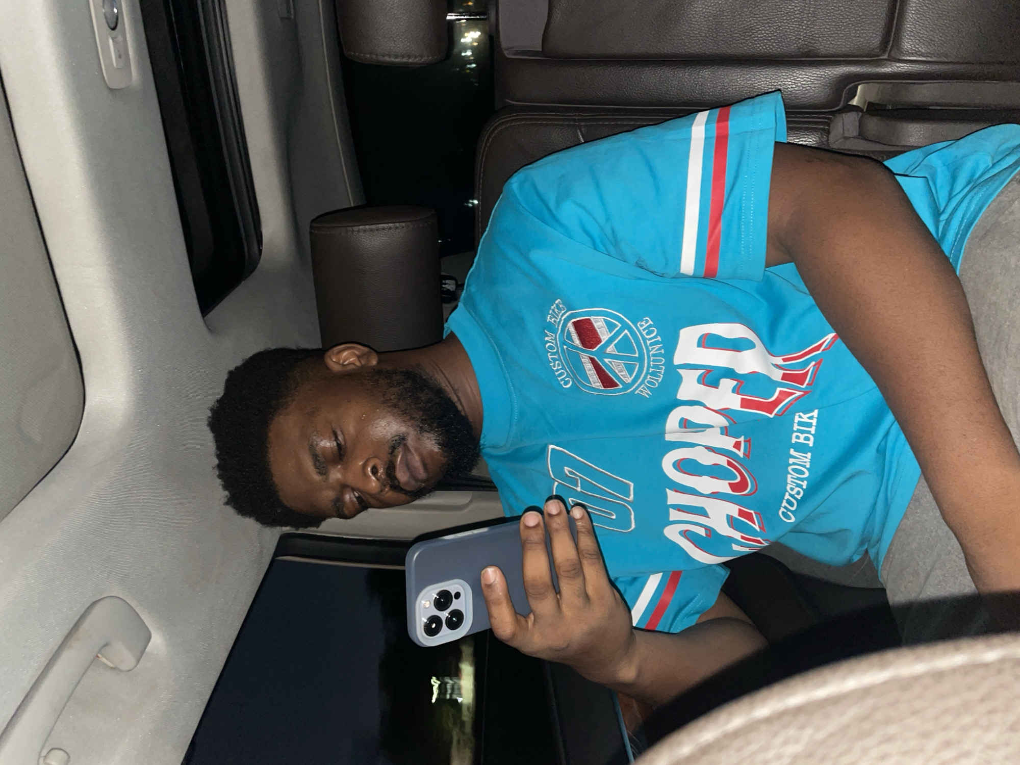 A medium shot shows a man with dark skin, short dark hair, and a beard, sleeping while holding a mobile phone in a car. He is wearing a light blue short-sleeved shirt with white and red stripes on the sleeves, and grey shorts. The text "CUSTOM WOL..." and "CHOPPER CUSTOM BIK" are visible on the shirt. The car's interior has grey roof upholstery, and the man is leaning against the interior. The car has dark brown leather seats. Outside the window, city lights are seen, suggesting a nighttime environment. The specific location of Jiru, Nigeria, cannot be determined from the image alone.