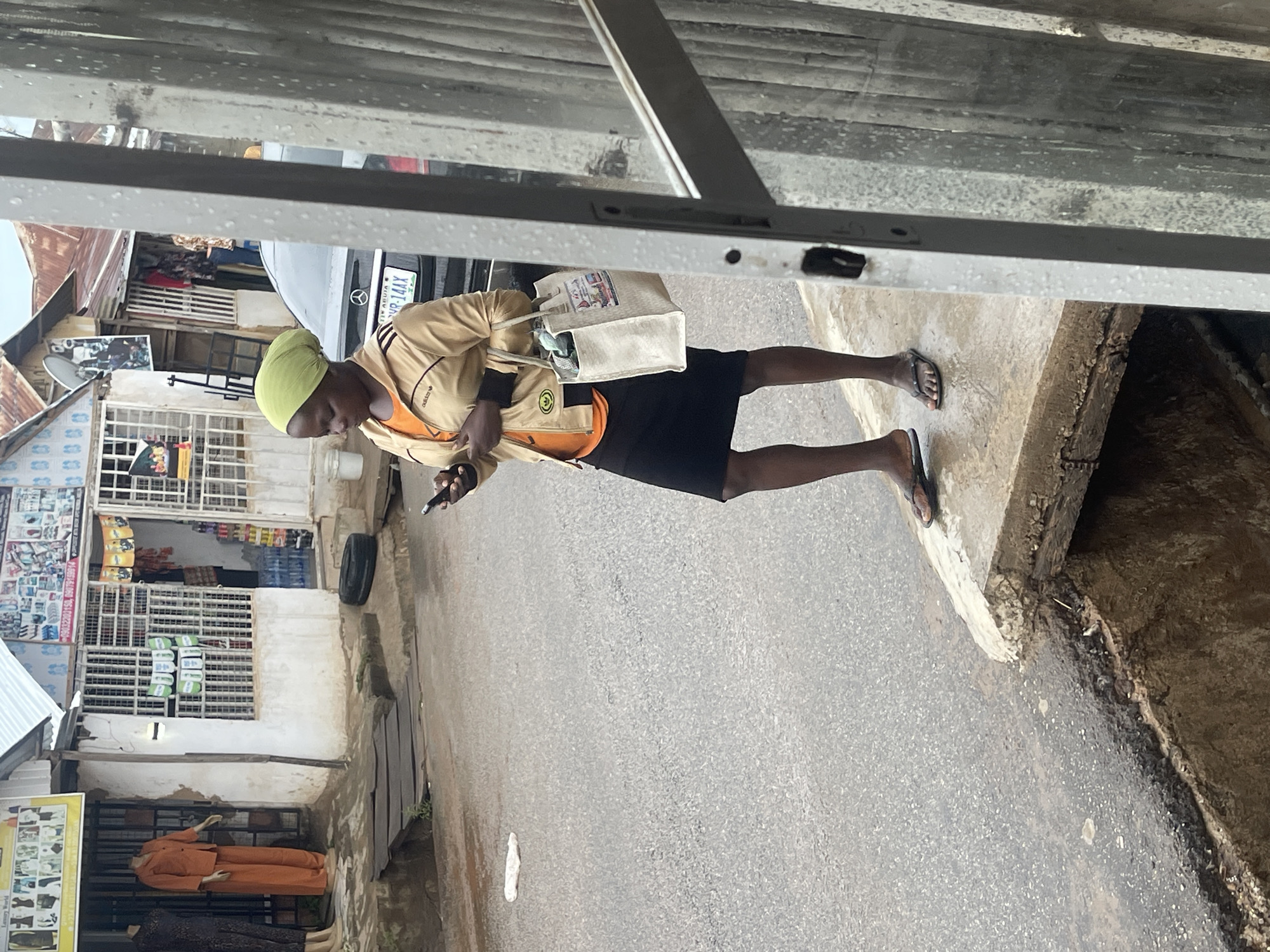 This candid street photograph, taken in Abuja, Nigeria, captures a person walking on an asphalt road next to a concrete curb and an open drain. The individual, appearing to be an adult with dark skin, is wearing a light green head covering, a khaki-colored jacket with black detailing and an Adidas logo over an orange shirt, and a black skirt. They are wearing black flip-flops. The person holds a mobile phone in their right hand and carries a light-colored reusable shopping bag over their left shoulder, appearing focused on their phone or the ground.
The background reveals a typical urban street scene in Abuja. Various buildings, likely shops or residences, line the street, some featuring corrugated metal roofs and barred windows. One visible shop displays clothing on mannequins, with signage partially legible as "LUXURY WORLD" and "FASHION." Another shop front shows packaged goods. A dark-colored car, possibly a Mercedes-Benz, is parked with its license plate clearly displaying "ABUJA VR-144X." The sky appears overcast, suggesting daytime. The foreground of the image is framed by a metallic structure, possibly an awning or part of a building's overhang. The overall scene depicts a routine moment in a lively commercial or residential area.