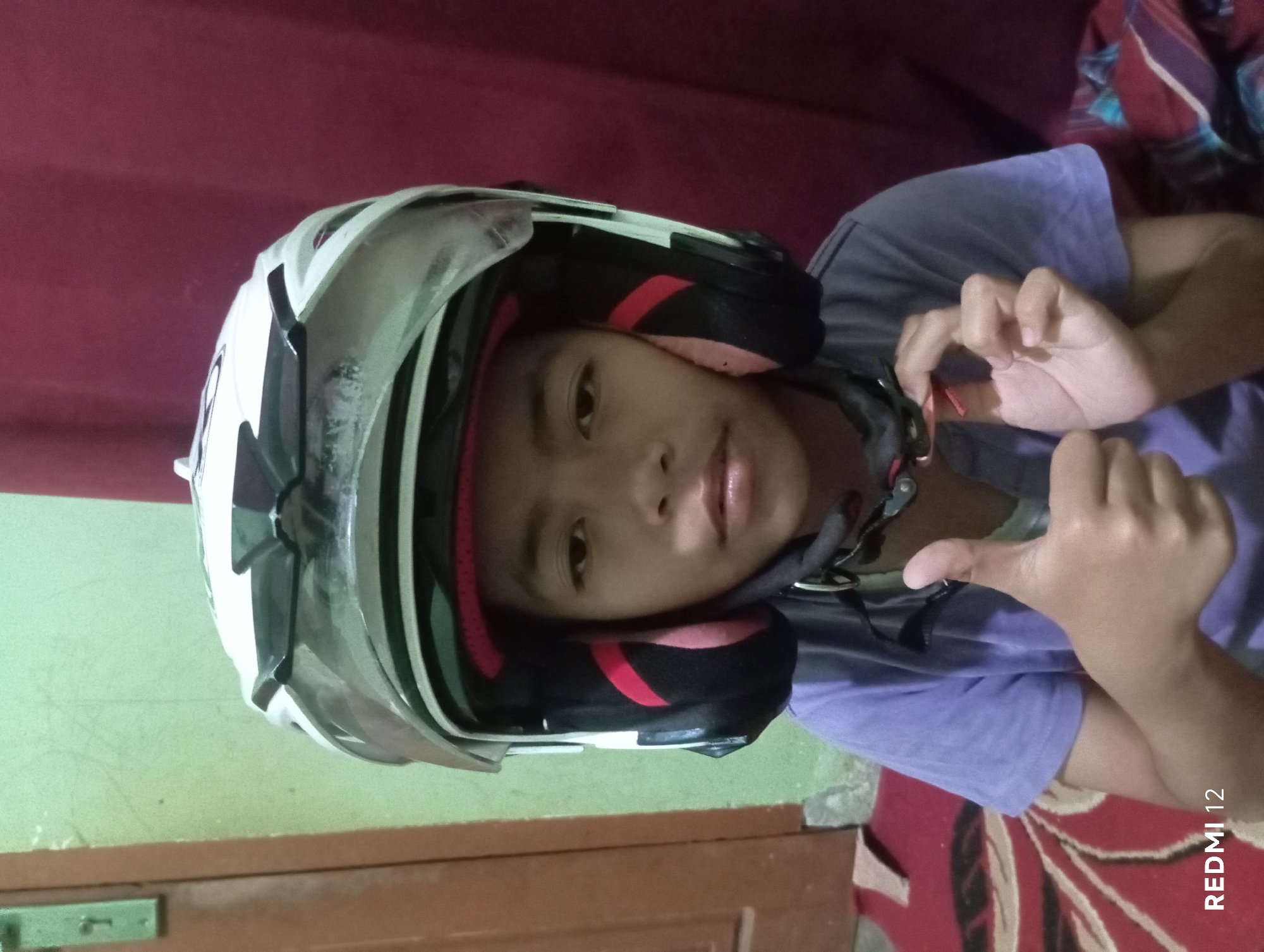 A young person is wearing a white and black helmet and a purple t-shirt, giving a thumbs up. The person's face is visible. They are indoors, with a light green wall behind them and a dark red curtain to the right. A portion of a wooden door is visible on the left. A patterned rug is on the floor. The image is tagged with "REDMI 12". This appears to be a casual, personal photograph taken in a residential setting, likely in Pagar Alam, Indonesia.