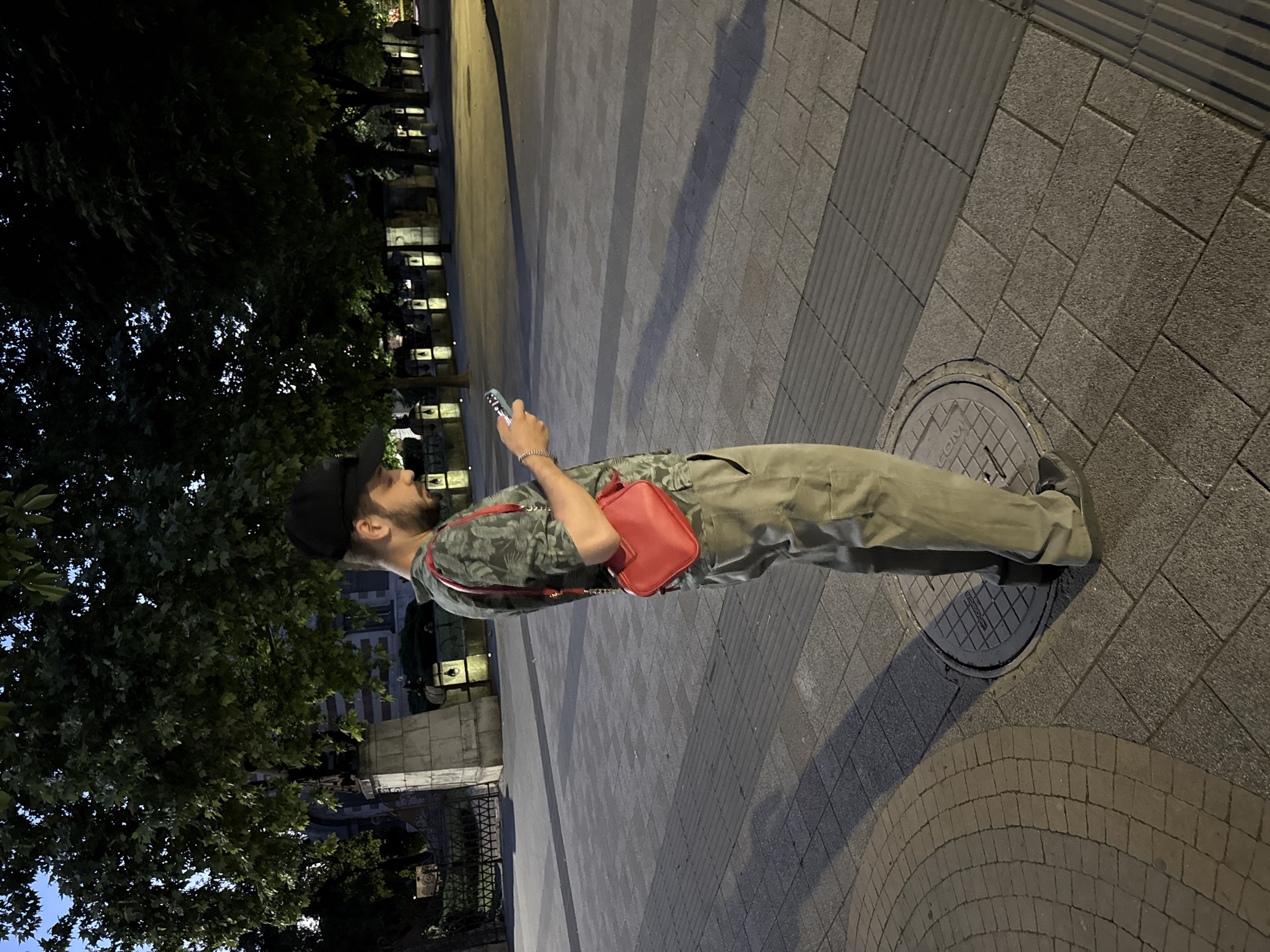 A man stands on a sidewalk in Pleven, Bulgaria, looking at his phone. He is wearing a camouflage t-shirt, khaki pants, and a black baseball cap. A small red purse is slung across his chest. The sidewalk is paved with grey and brown rectangular tiles, with a circular metal manhole cover visible near his feet. In the background, trees with dark green foliage line a path, and streetlights cast warm glows. A building with stone accents and decorative lanterns is partially visible behind the trees. The sky above is dark, suggesting it is evening or night.