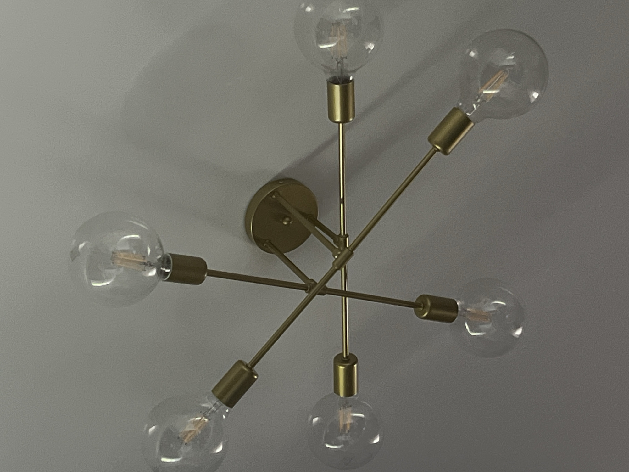 This is a close-up, low-angle shot of a modern, multi-bulb light fixture mounted on a grey wall. The fixture is made of brushed gold-colored metal and has a central mounting plate. Five exposed Edison-style incandescent bulbs are attached to thin, gold-colored arms that radiate outwards from the center. The bulbs are clear glass and each contains a visible filament. The lighting appears to be dim, as the bulbs are not illuminated, casting soft shadows on the wall. The overall aesthetic is minimalist and contemporary. There is no visible text. The image was likely taken indoors.