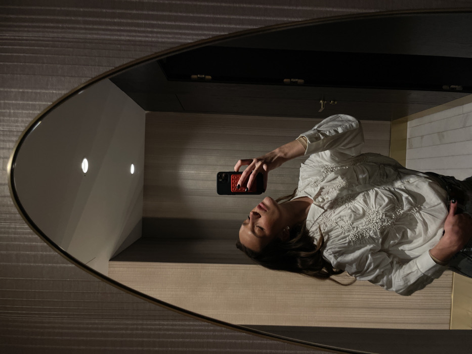 A woman reflected in an oval mirror takes a selfie with her phone. The woman, wearing a textured white shirt, holds a black phone with a red case. The background includes a textured wall, a lit, white wall corner, and dark cabinetry. The mirror itself has a thin gold trim and reflects shadows, adding depth to the image.