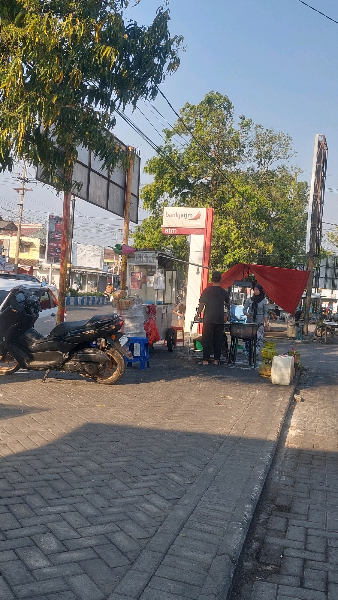 A medium shot captures a street scene in Nologaten, Indonesia, on a clear day. In the foreground, a dark-colored scooter is parked on a herringbone-patterned brick sidewalk. Beyond the scooter, a street vendor's cart, likely selling food, is set up. Two individuals are interacting near the cart; a man in a black t-shirt and dark pants stands, and a woman wearing a headscarf and a grey top with patterned pants is seated. There are multiple gas tanks and cooking equipment present. A red canopy shelters the area where the people are. Behind the vendor's stall, a prominent "bankjatim ATM" sign is visible on a white and red pillar, surrounded by trees. The background shows more trees, buildings, and utility poles, indicating an urban environment. Sunlight casts shadows on the brick pavement.